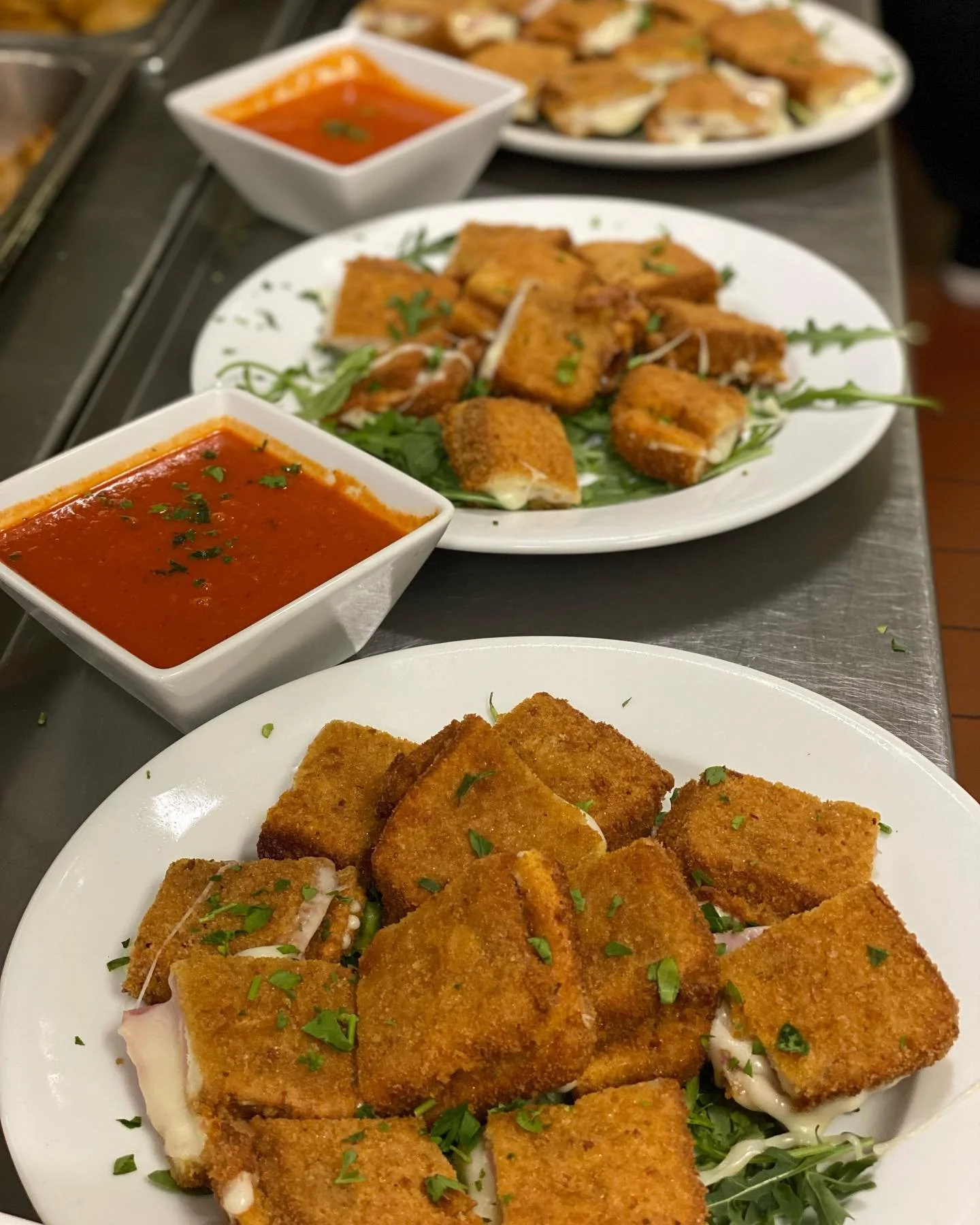 Several plates of breaded and fried cheese sticks garnished with chopped herbs, along with two bowls of marinara sauce, are arranged on a metal countertop.