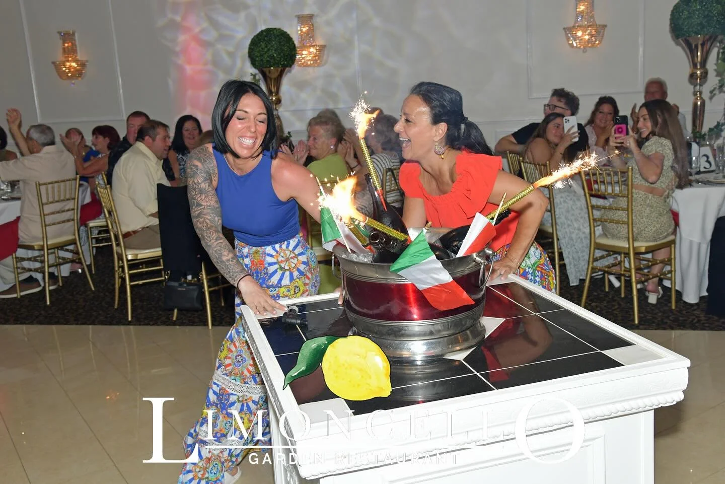 Two women celebrating with a cake and sparklers at a birthday party, with guests seated at tables in the background.