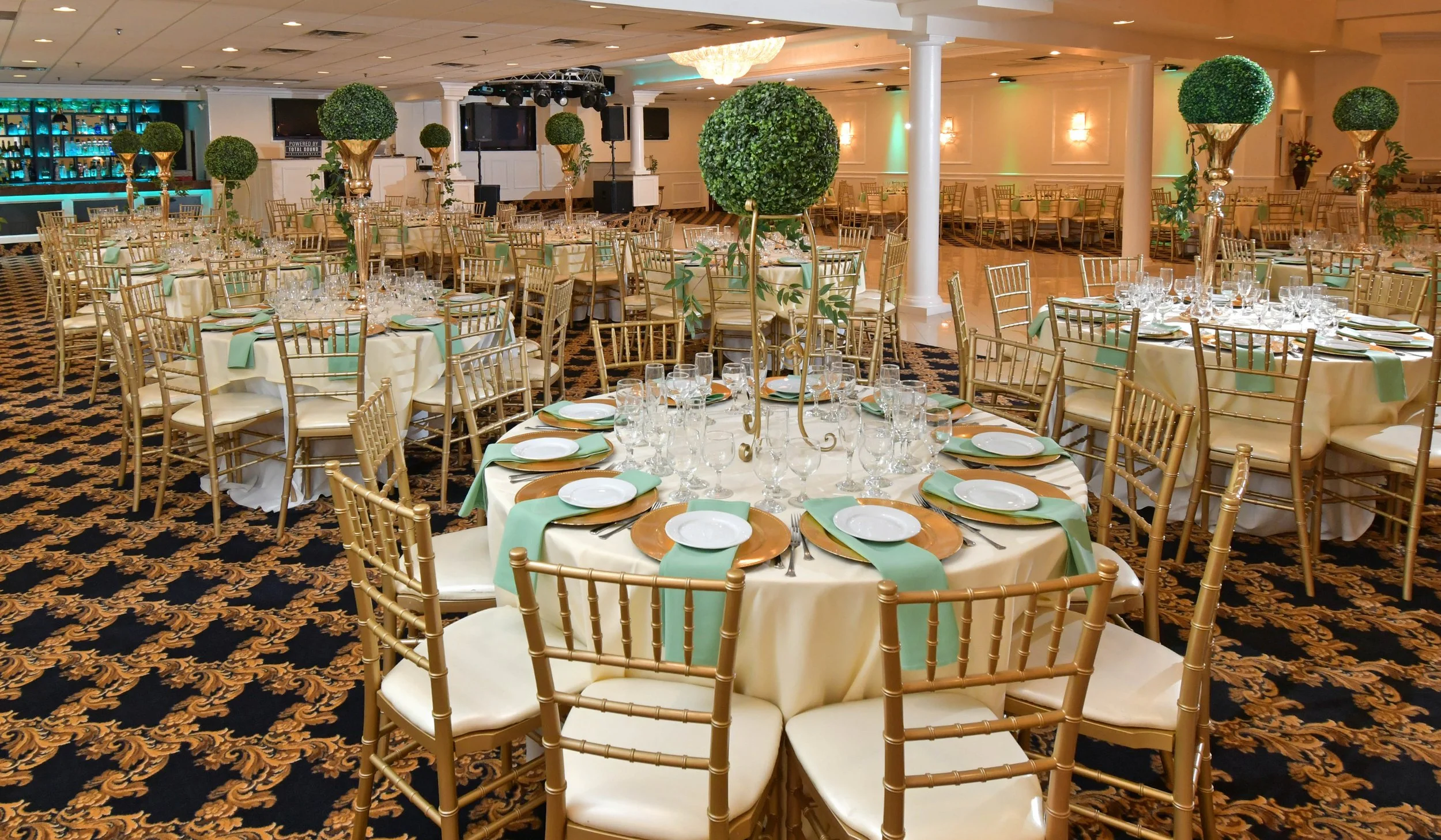 Elegant banquet hall with round tables decorated with cream tablecloths, gold chairs, and tall centerpieces of green topiary balls in gold stands. The tables are set with glassware, plates, and green napkins, and a bar is visible in the background.