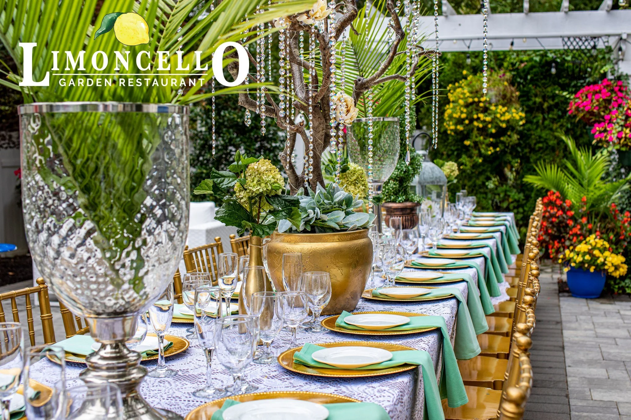 A beautifully decorated outdoor dining table at Lemoncello Garden Restaurant with floral centerpieces, crystal glassware, and colorful table settings surrounded by lush greenery and blooming flowers.