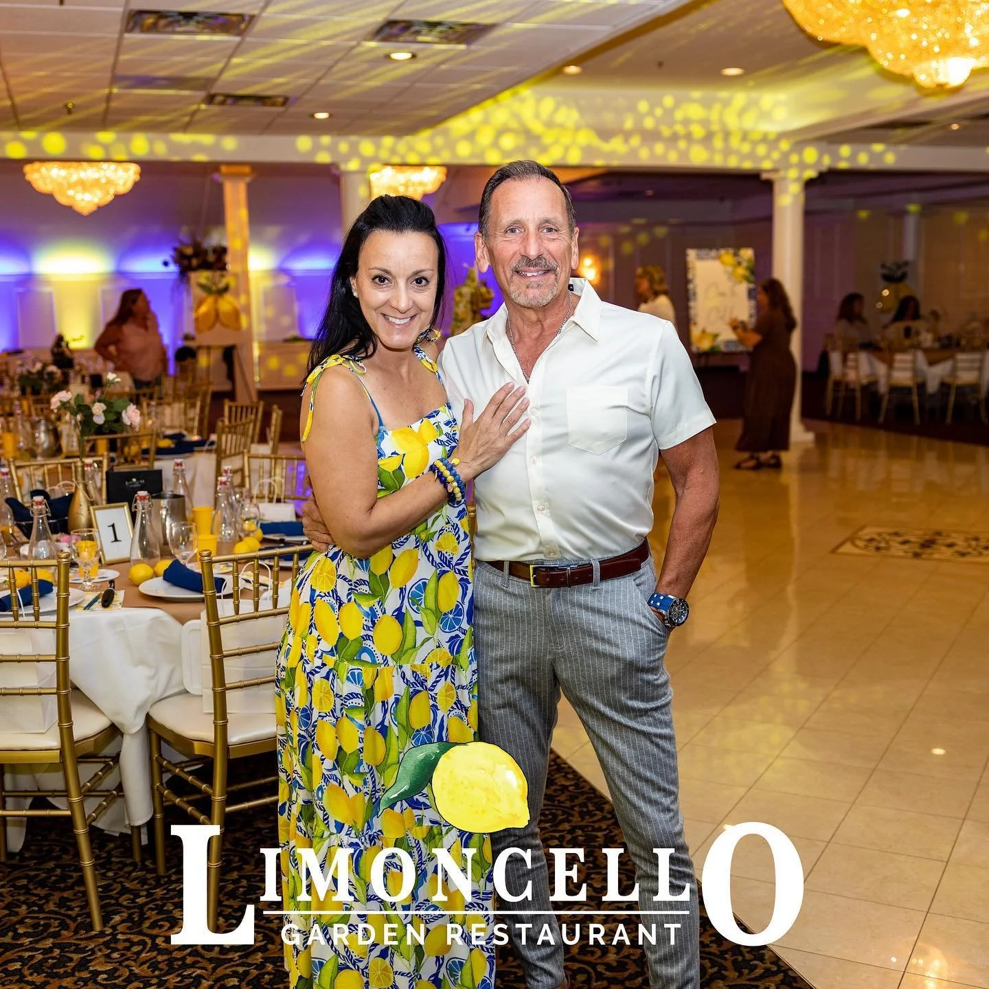 A smiling woman in a lemon-patterned dress and a man in a white shirt and striped pants stand together at a decorated event venue, with tables set for a celebration and a sign reading 'Limoncello Garden Restaurant' in the foreground.