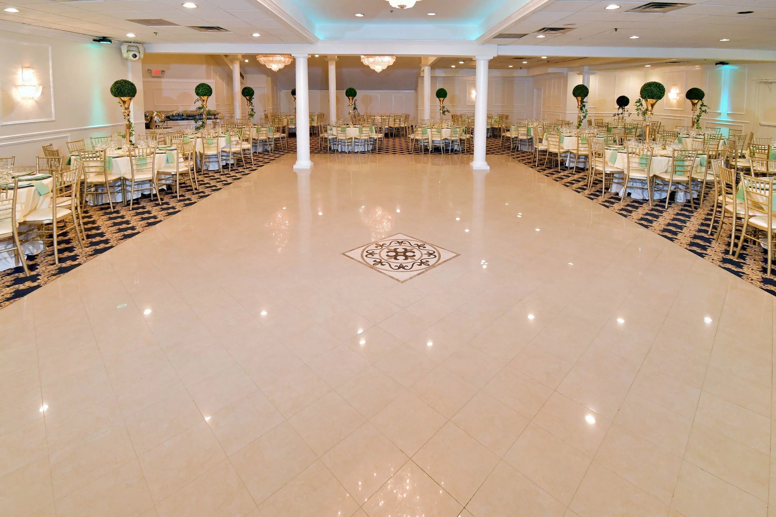 Elegant banquet hall with round tables set with white tablecloths, gold chairs, and floral centerpieces. Decor includes white walls, chandeliers, and columns, with a polished tile floor and a decorative pattern in the center of the floor.