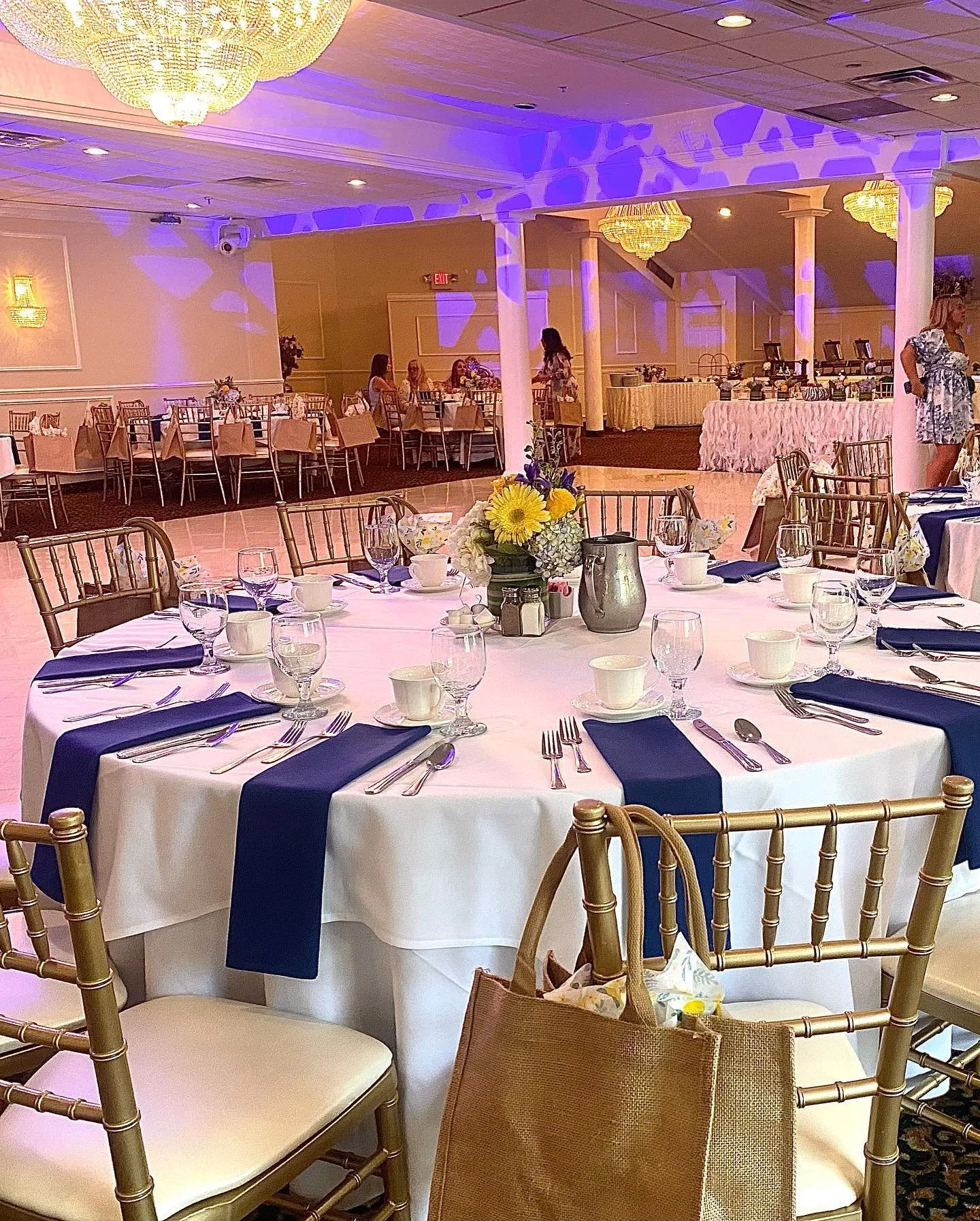 Wedding reception hall with round tables decorated with blue napkins, white tablecloths, and floral centerpieces. There are gold chairs around the tables, and a buffet table against the wall. Purple and yellow lighting create a festive atmosphere.