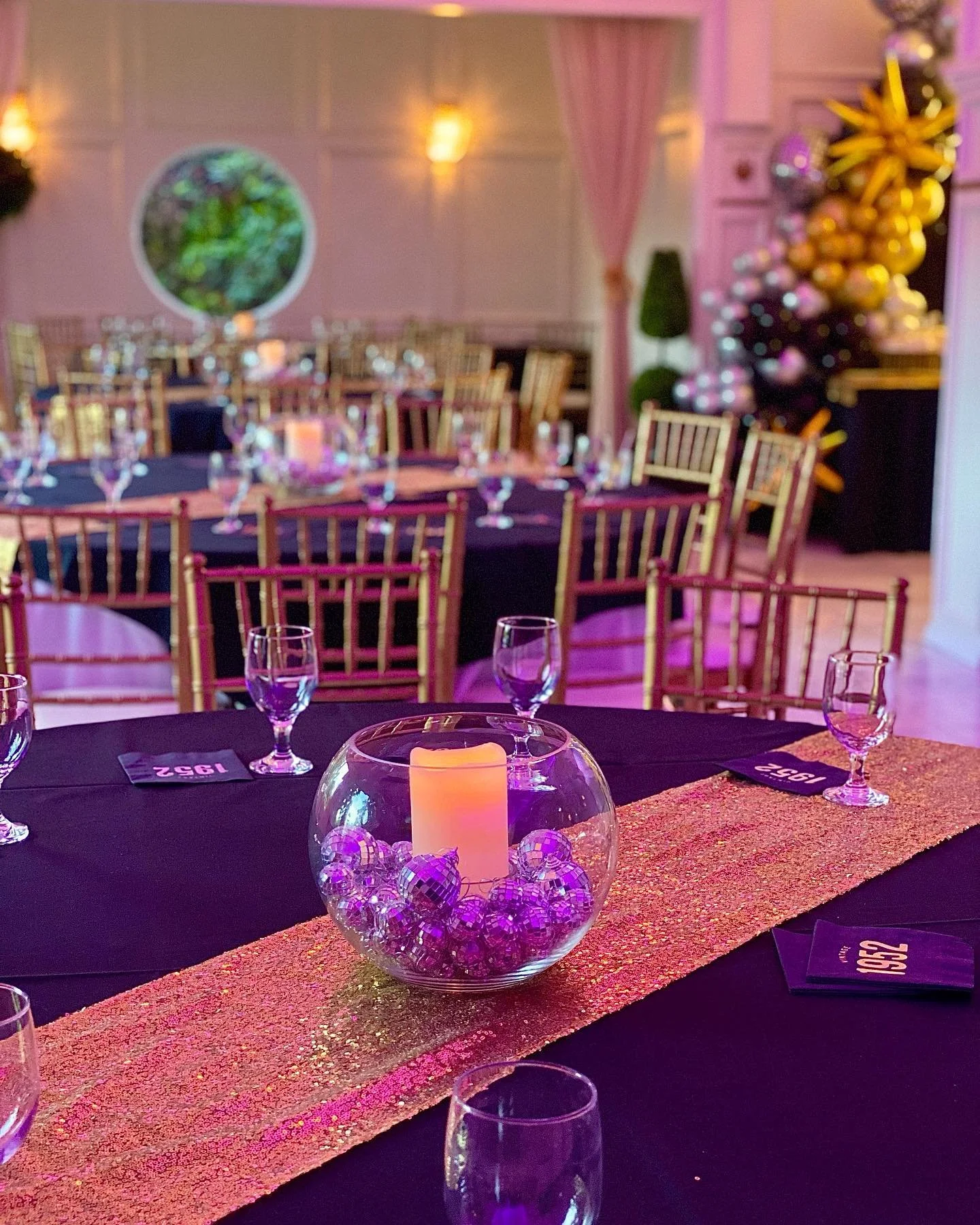 Holiday party setup with tables decorated with black tablecloths, gold chairs, and a gold and pink glittery table runner. Centered centerpiece features a candle inside a glass bowl filled with purple and silver disco balls. In the background, there i