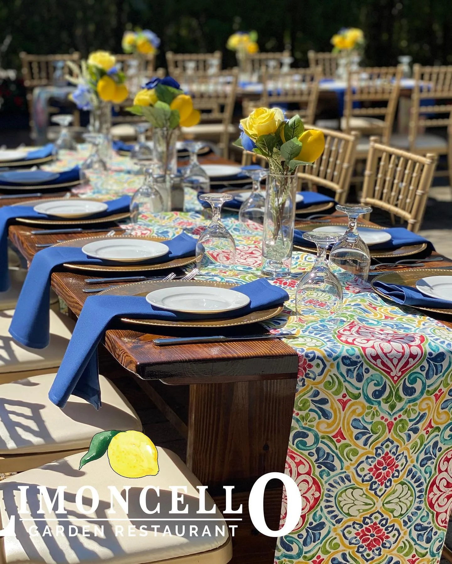 Decorated outdoor dining table with colorful tablecloth, blue napkins, white plates, upside-down wine glasses, and yellow and blue flowers in tall vases at Le Moncel Garden Restaurant.