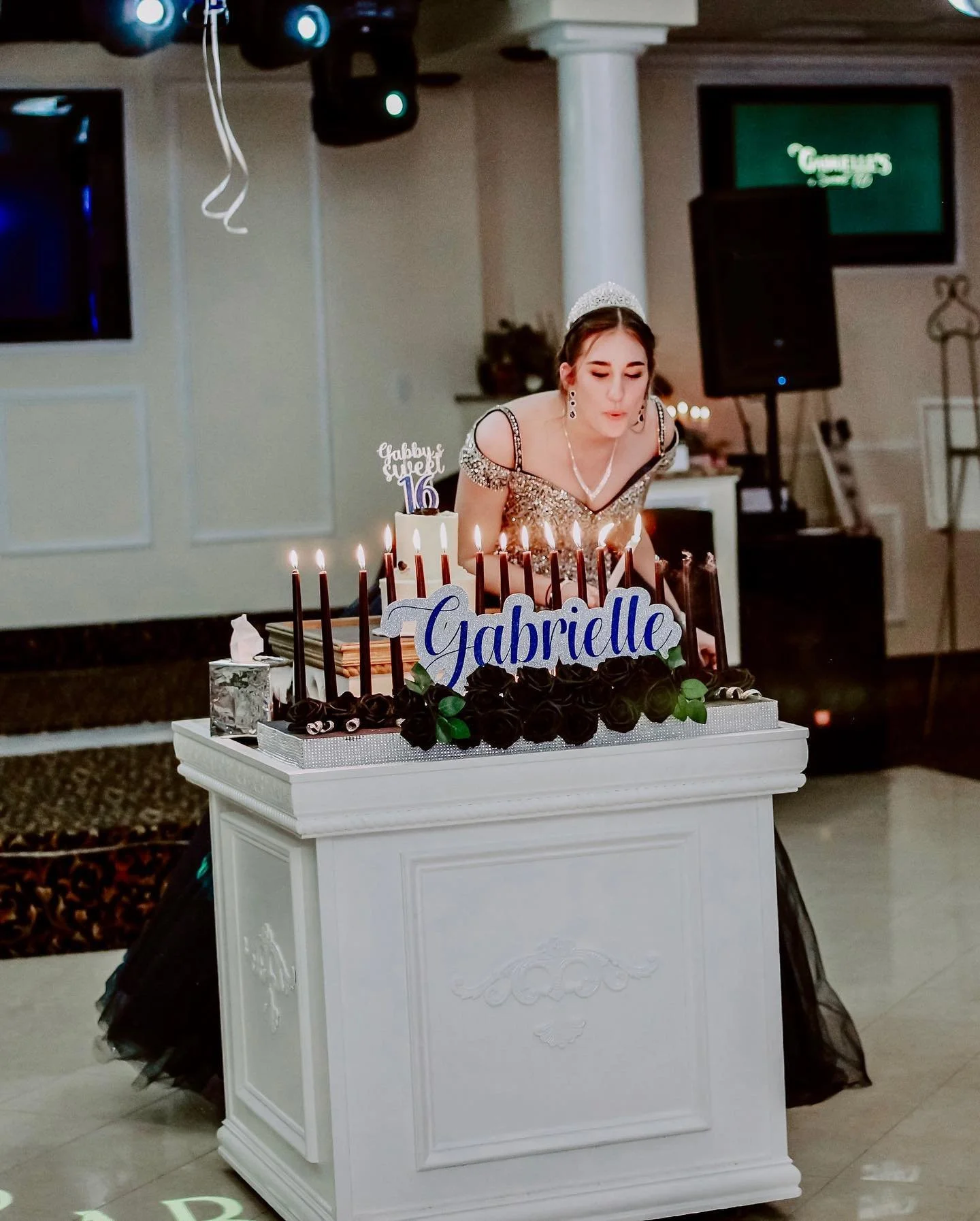 Young woman in a sparkling dress and tiara blowing out candles on a birthday cake decorated with black roses and a blue 'Gabrielle' sign, celebrating her 16th birthday.