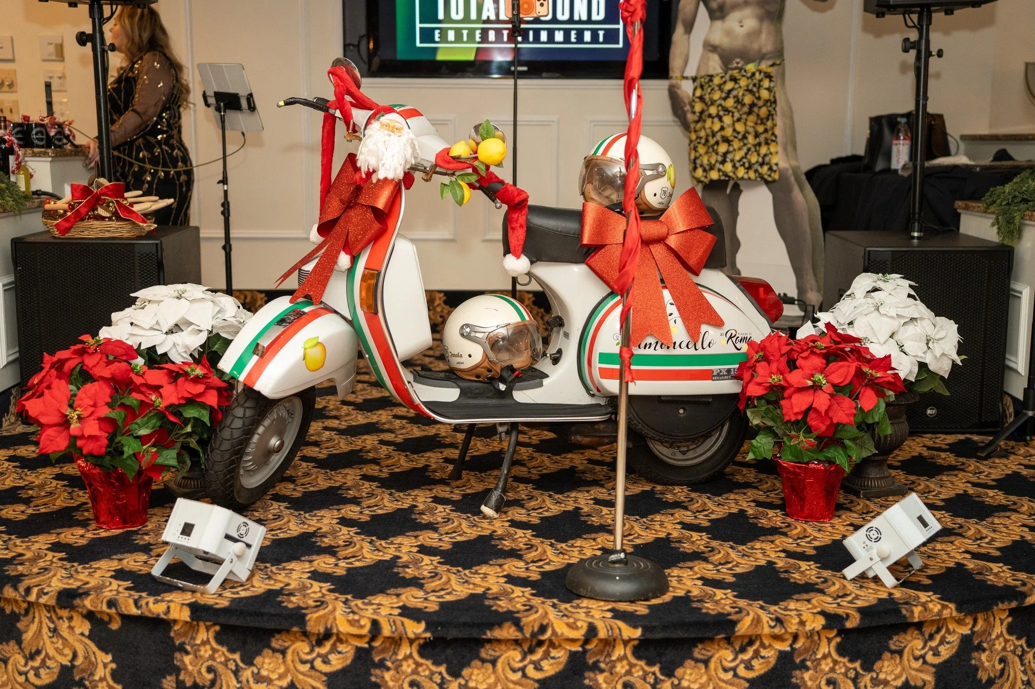 A decorated vintage scooter with Christmas decorations, including a Santa figurine and red bows, surrounded by poinsettia plants on a patterned carpet, with people and equipment in the background.