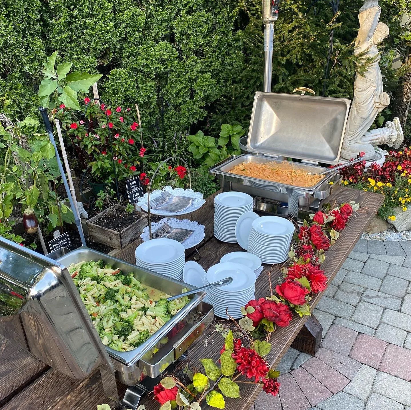 Outdoor buffet table with pasta, salad, and plates set among flowers and greenery.