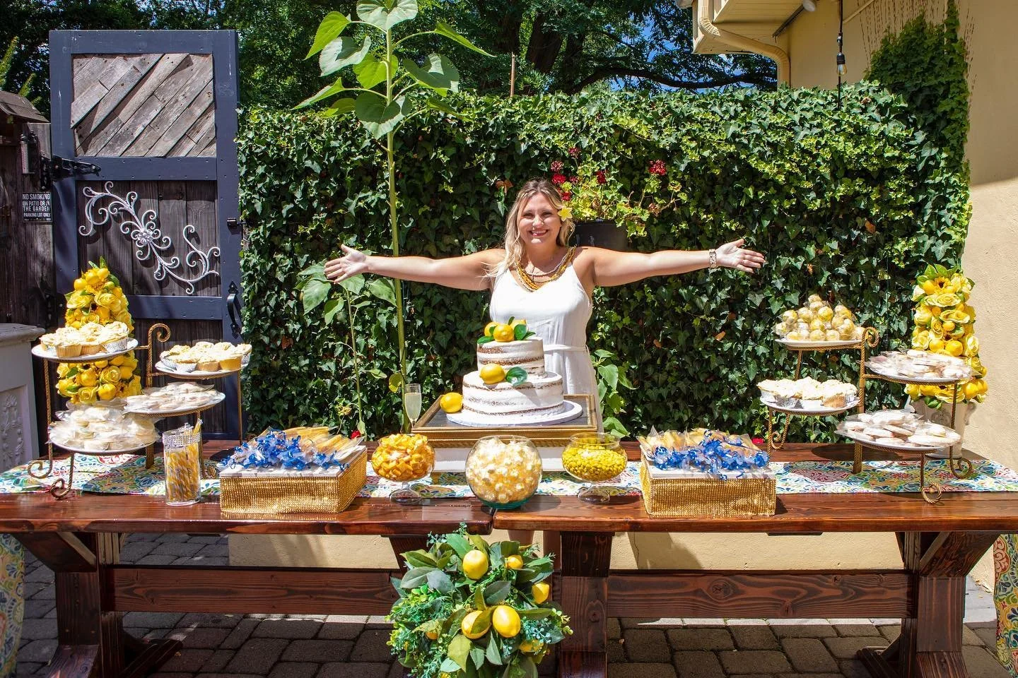 A woman celebrating with her arms outstretched behind a dessert table decorated in yellow and lemon-themed decorations. The table has a tiered lemon cake, lemon candies, and flowers, set outdoors against a green vine-covered wall.