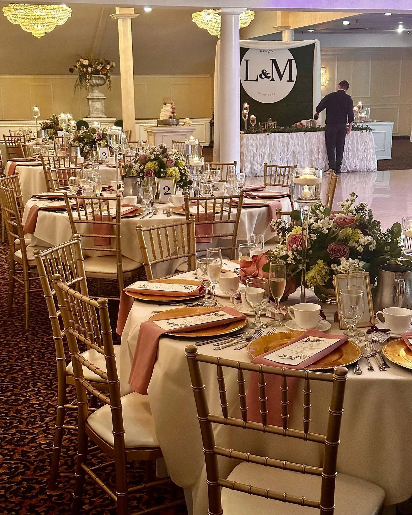 Wedding reception with round tables decorated with pink and white floral centerpieces, gold chairs, pink napkins, and a dessert table in the background with wedding cake and decorations.