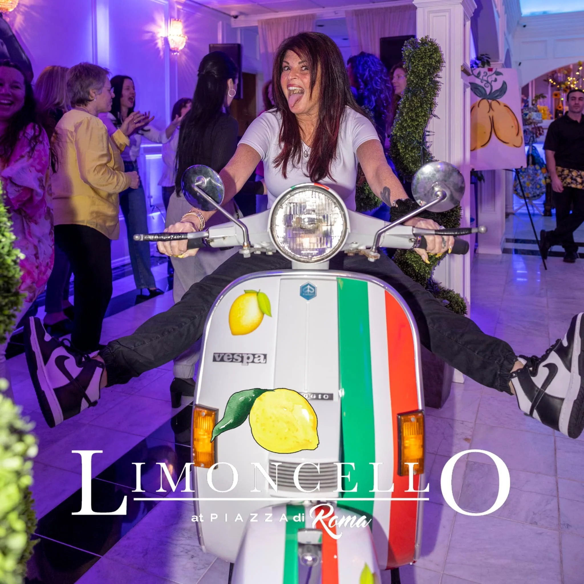 A woman with brown hair and a white T-shirt sitting on a Vespa scooter decorated with lemon images and the Italian flag colors, surrounded by people at an indoor event.