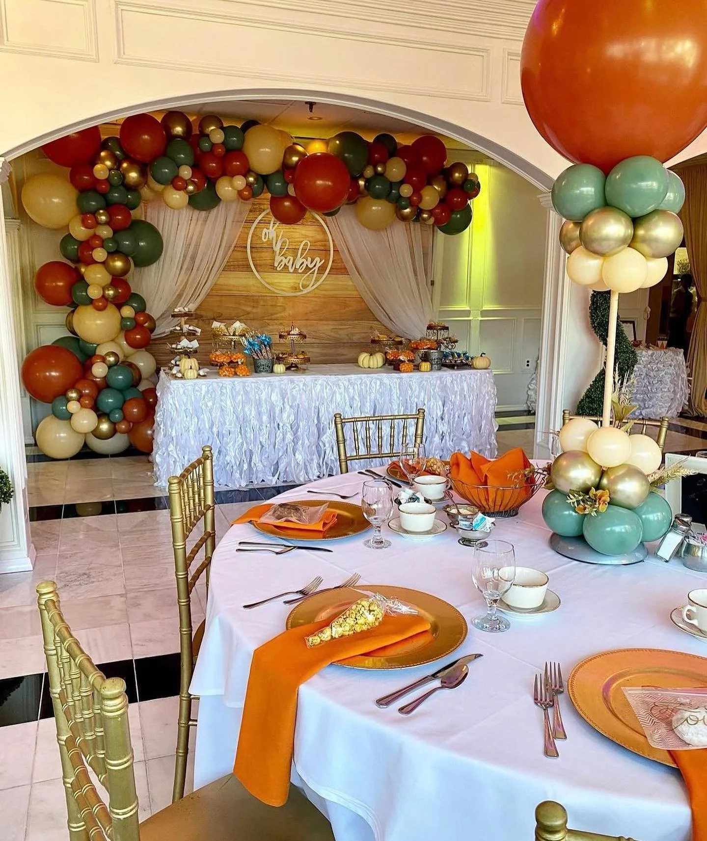 Decorated event space for a celebration with a balloon arch, a table with orange napkins, and a dessert table with a 'Oh Baby' sign in the background.