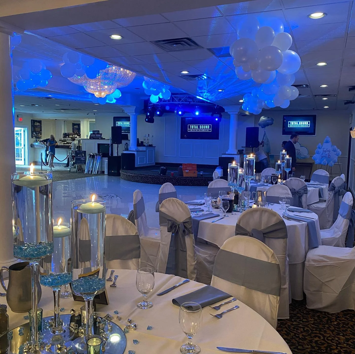 Banquet hall decorated with white and gray themed table settings, white chairs with gray sashes, candles on tall holders, balloons, and a stage with DJ equipment under blue lighting.