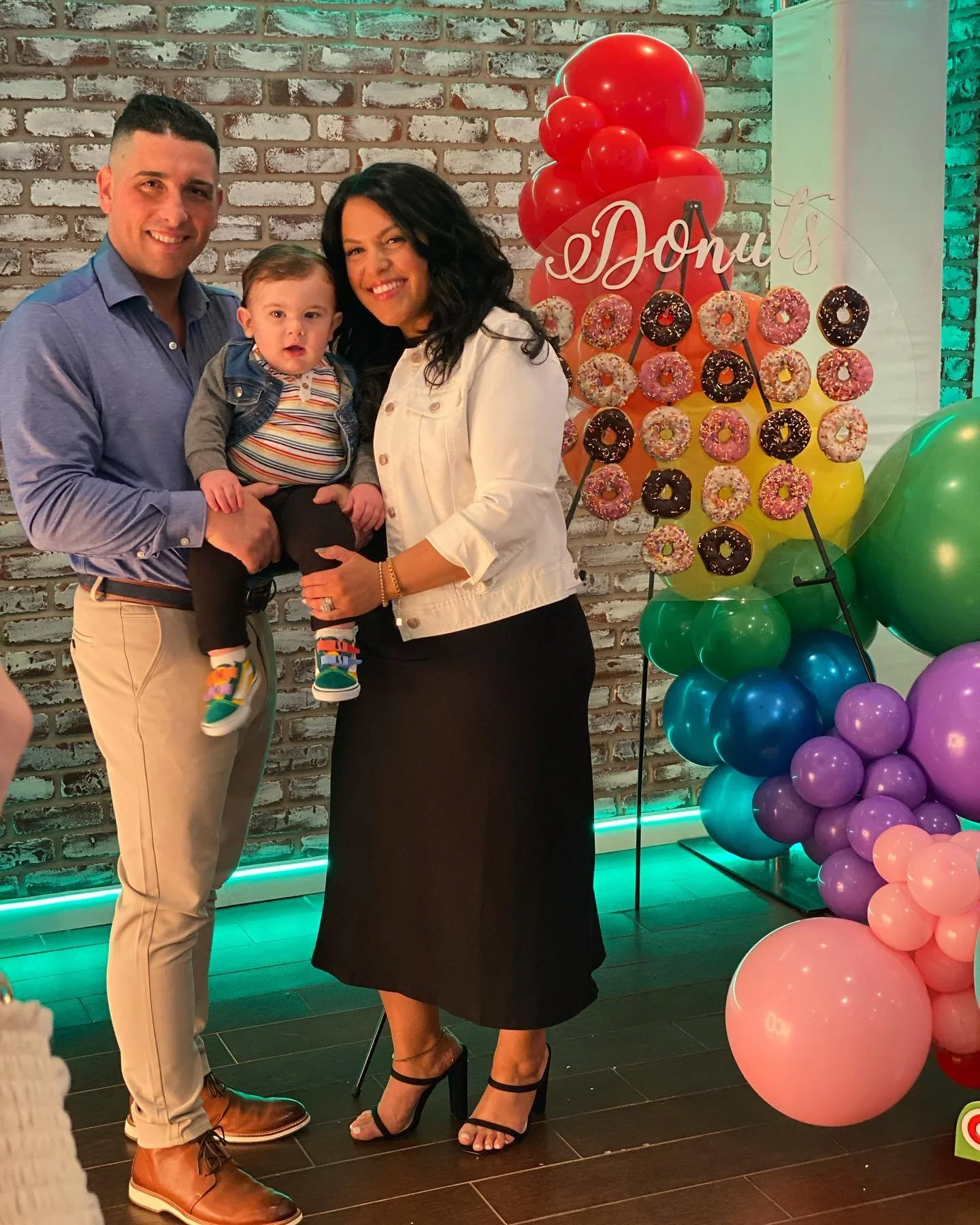 A family of three standing together at a birthday celebration with colorful balloon decorations and a donut display in the background.