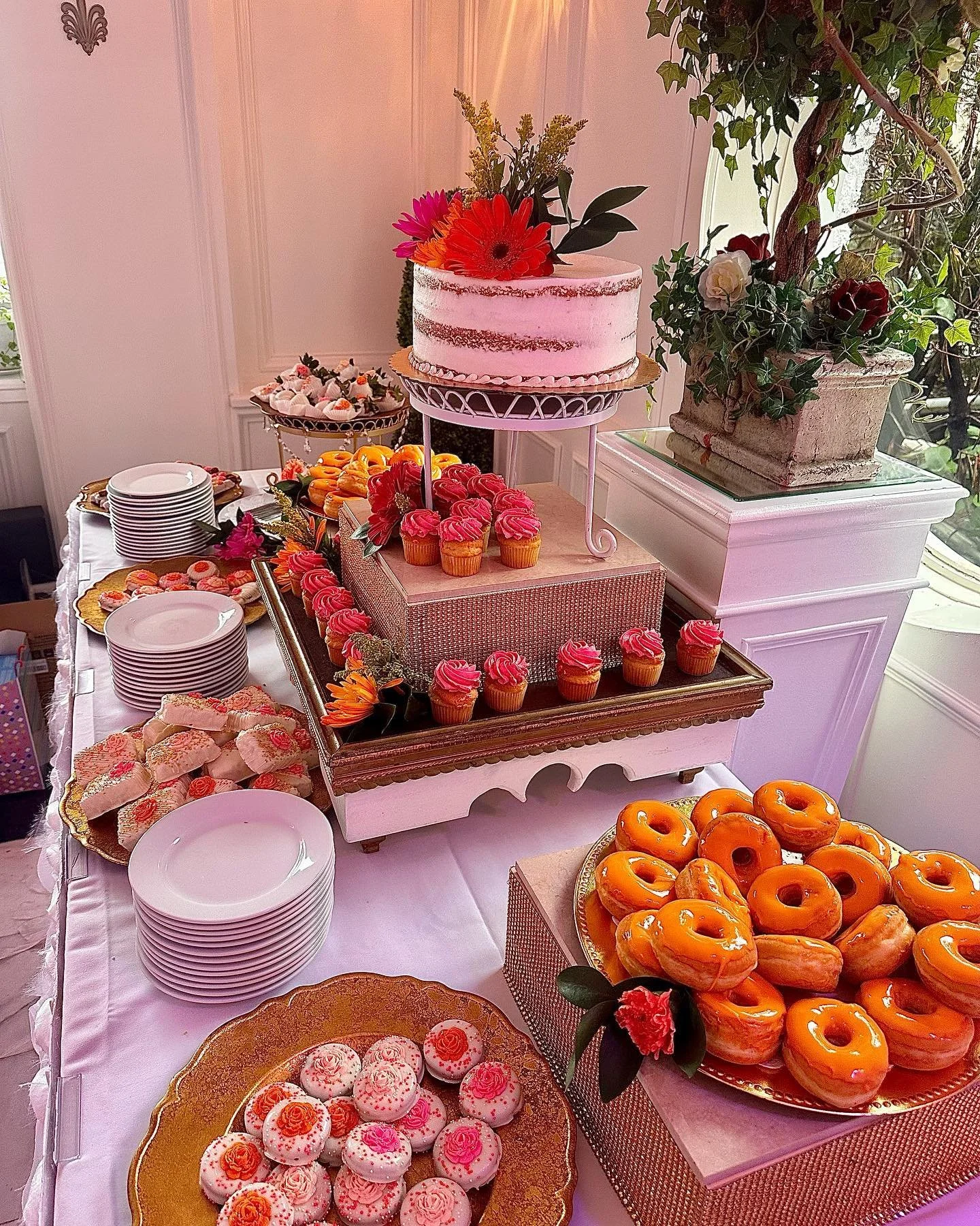 Dessert table with a pink layered cake decorated with flowers, surrounded by cupcakes, donuts, and cookies, all on elegant trays. Stacked plates and a flower arrangement are also visible.