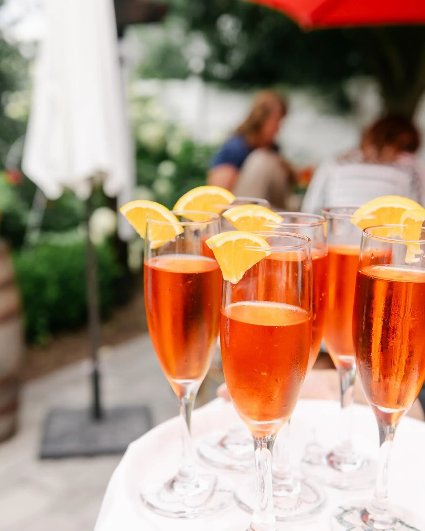 Seven glasses of pink sparkling drink with lemon wedges on the rim, set on a white table outdoors.