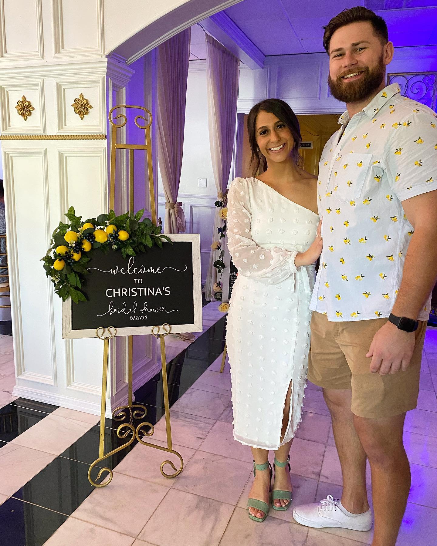 A smiling woman and man at a bridal shower celebration, standing beside a decorative welcome sign with lemon and greenery accents.