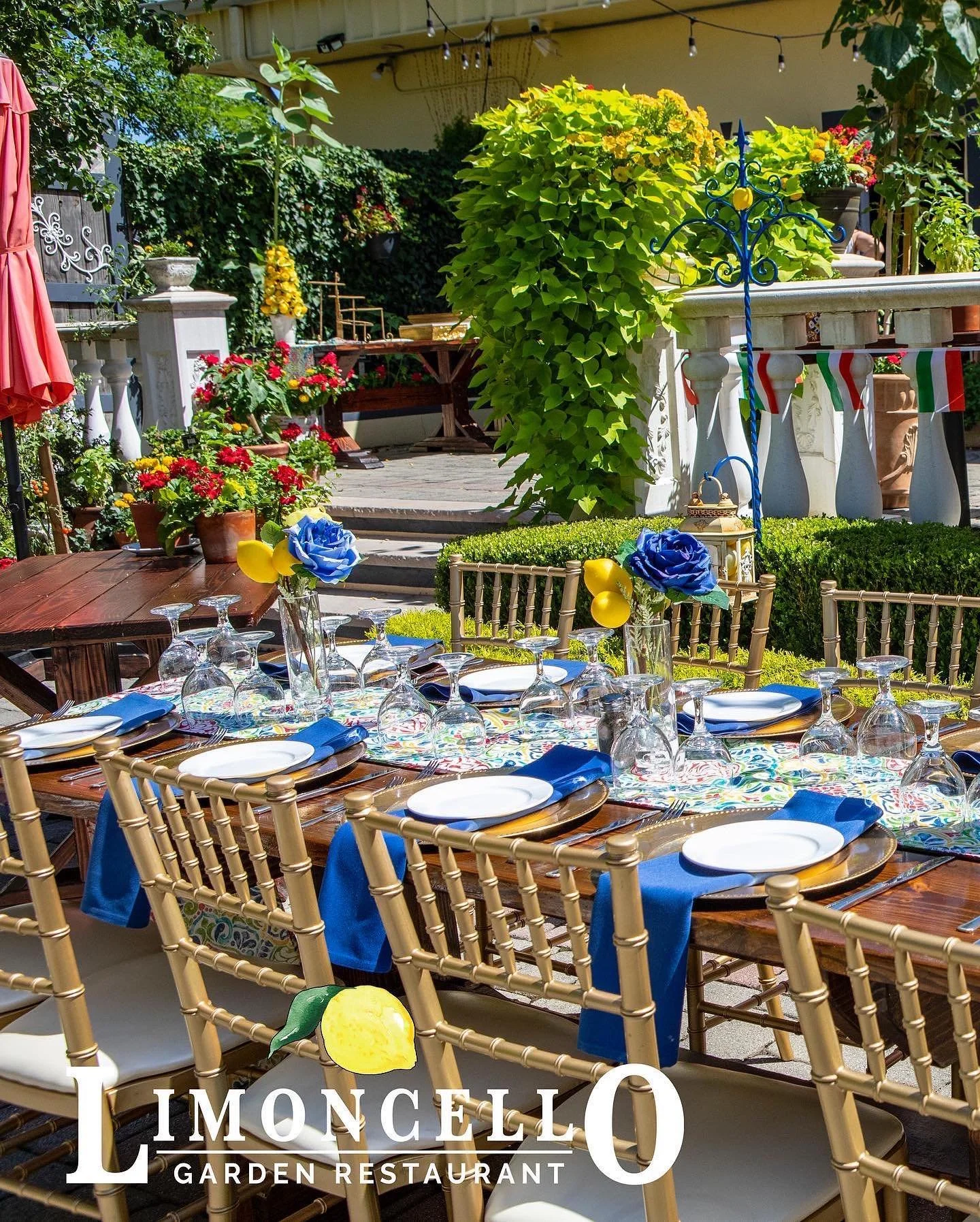 Outdoor garden restaurant with tables set for dining, decorated with blue and yellow flowers and balloons, surrounded by lush green plants, string lights, and patio furniture.