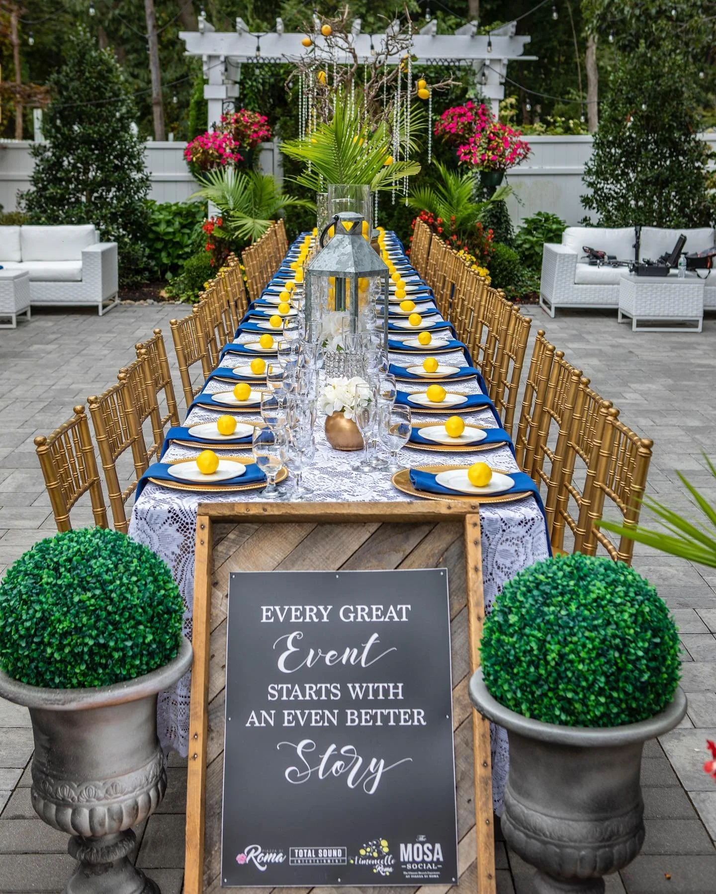 Outdoor wedding or event table setup with a long table, gold chairs, white tablecloth, and yellow decorations. A blackboard sign in front states, "Every great event starts with an even better story." Potted green plants flank the sign, and colorful f