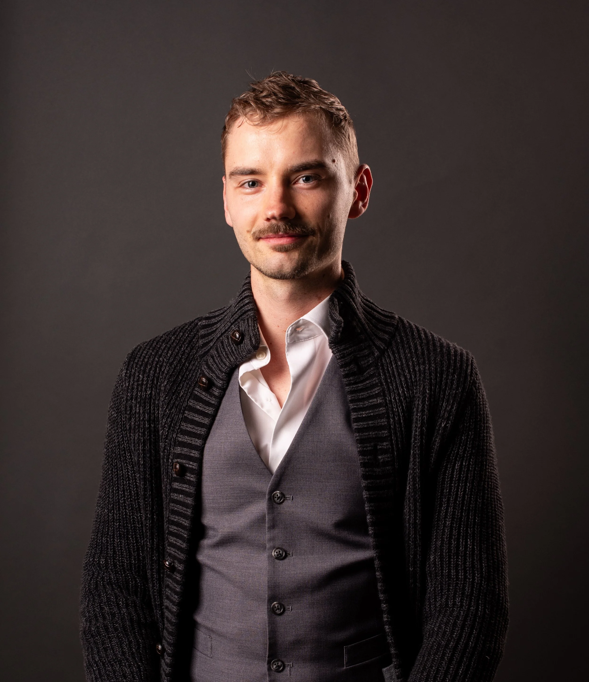 Portrait of a young man with short hair and a goatee, wearing a beige shirt, a dark cardigan, and a long beaded necklace with a tassel, smiling against a black background.