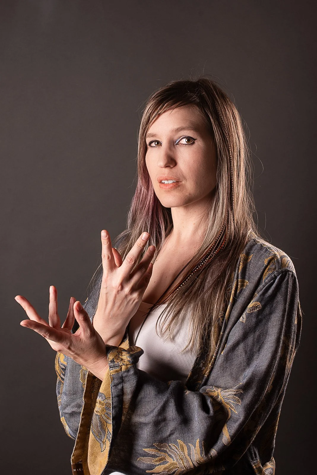 A woman with wavy brown hair and green eyes makes a gesture with her hands, wearing a black top and striped pants against a black background, illuminated with pink and red lighting.