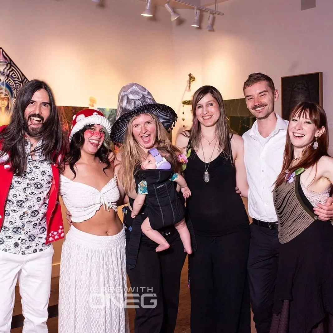 Group of eleven people posing playfully in an art gallery, with various artworks on the wall behind them and a large, colorful, textured circular artwork above. Some are making funny faces and gestures, with one person sitting on a red velvet couch.
