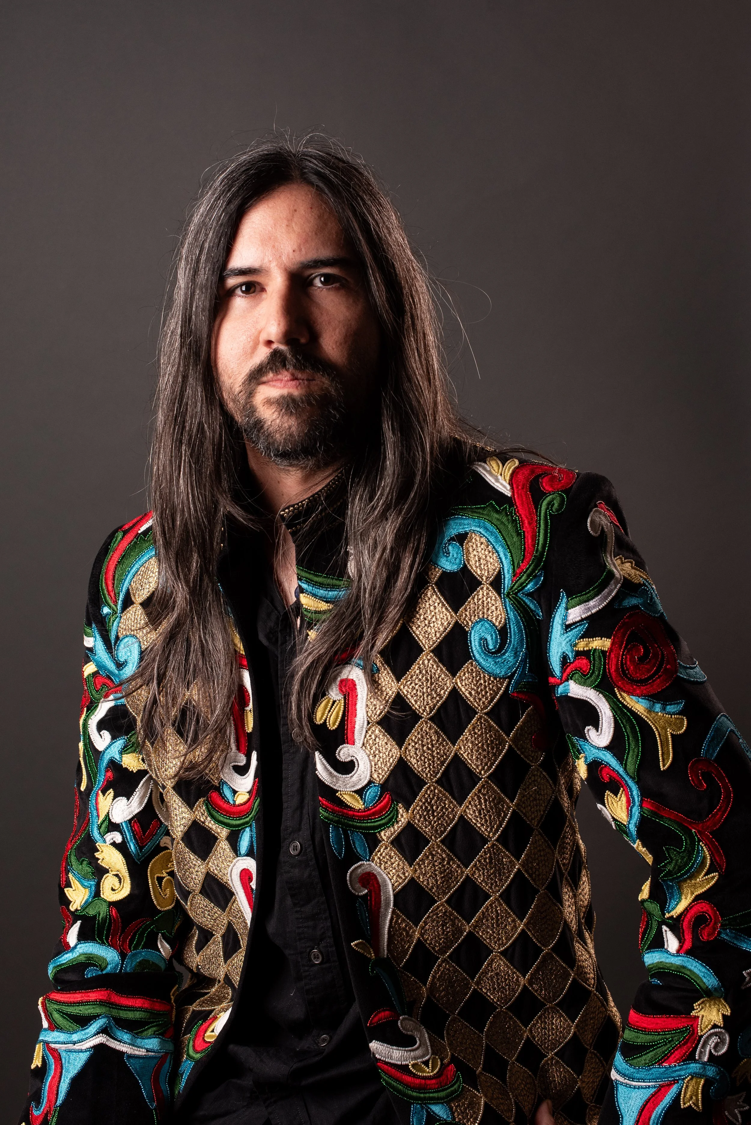 A man with long dark hair and a beard, wearing a colorful, ornate embroidered jacket, holding a ring to his chin, against a black background.