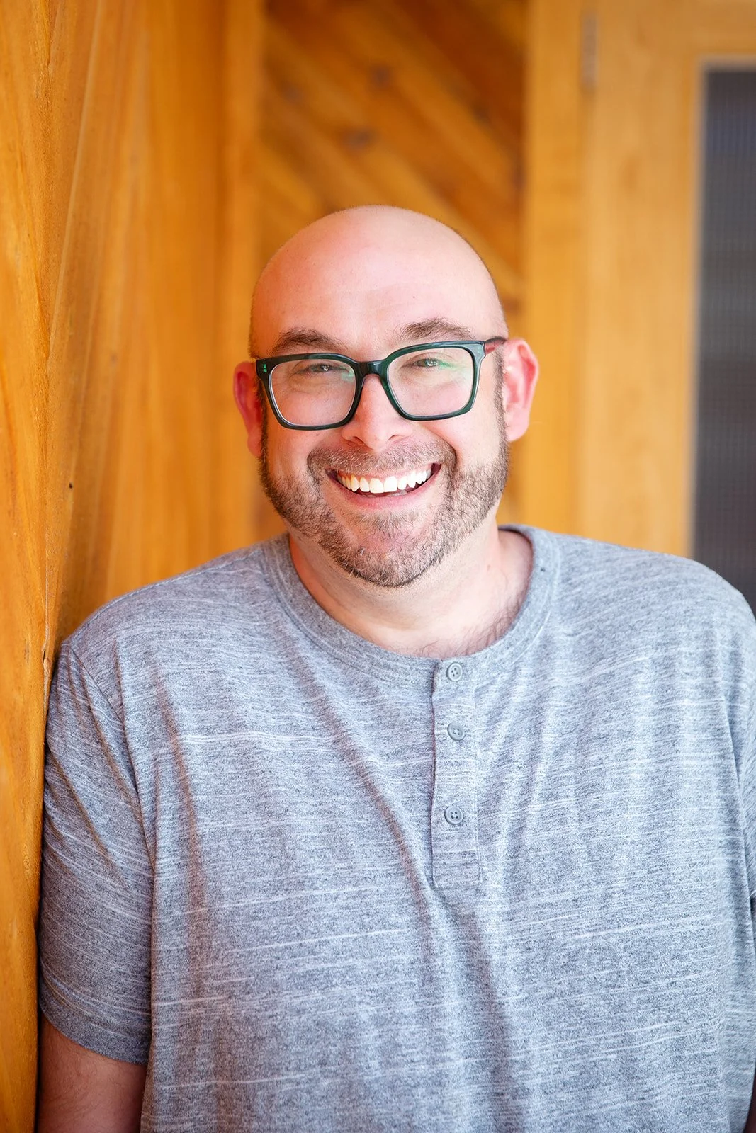 A smiling man with glasses and a bald head standing with arms crossed in front of a blue wall.