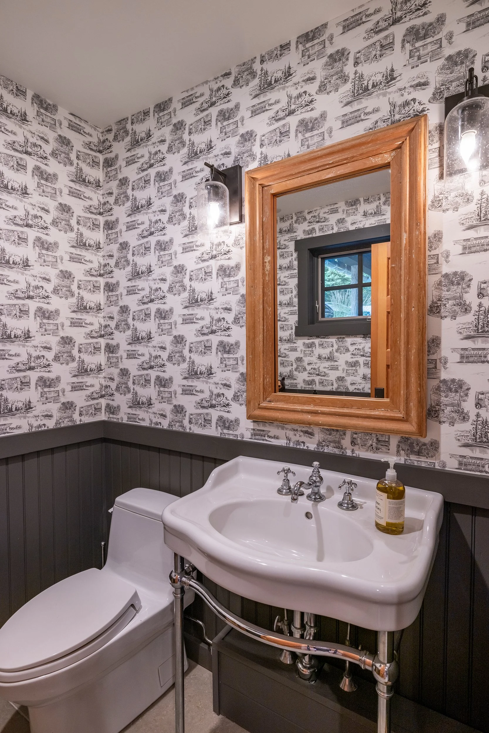 Small powder room with black and white wallpaper, a white toilet, a white vintage-style sink with chrome pipes, a wooden framed mirror, black wall-mounted lights, and a small window.