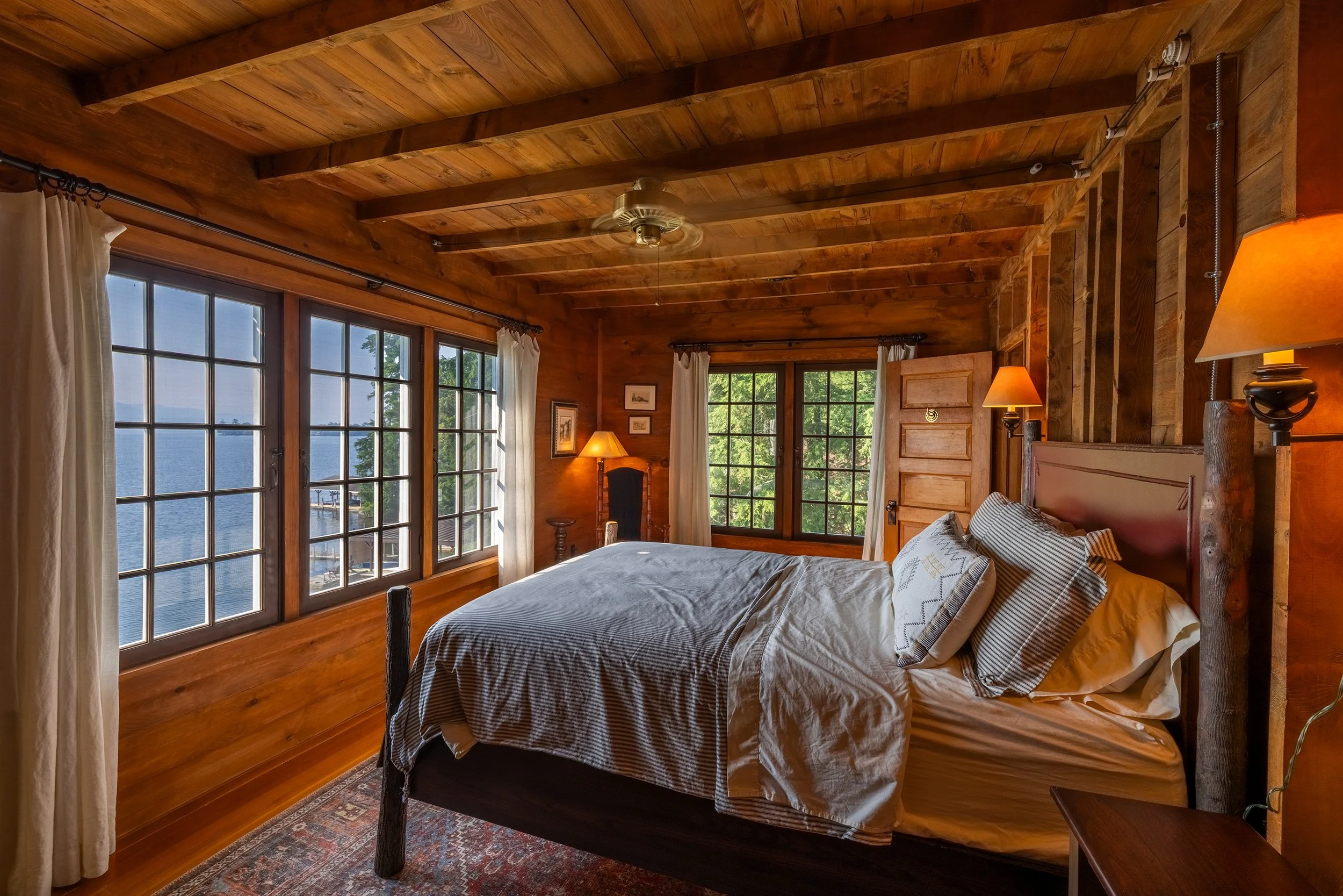 A cozy, rustic bedroom with wooden walls, ceiling, and furniture, featuring a large bed with striped pillows, surrounded by big windows overlooking a lake, with curtains and warm lighting.