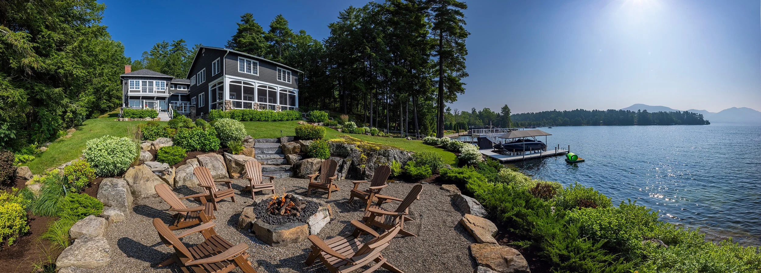 A lakeside house with a screened porch, surrounded by greenery, rocks, and a fire pit area with wooden chairs. The house overlooks a calm lake with docked boats and a pier, with mountains in the distance under a clear blue sky.