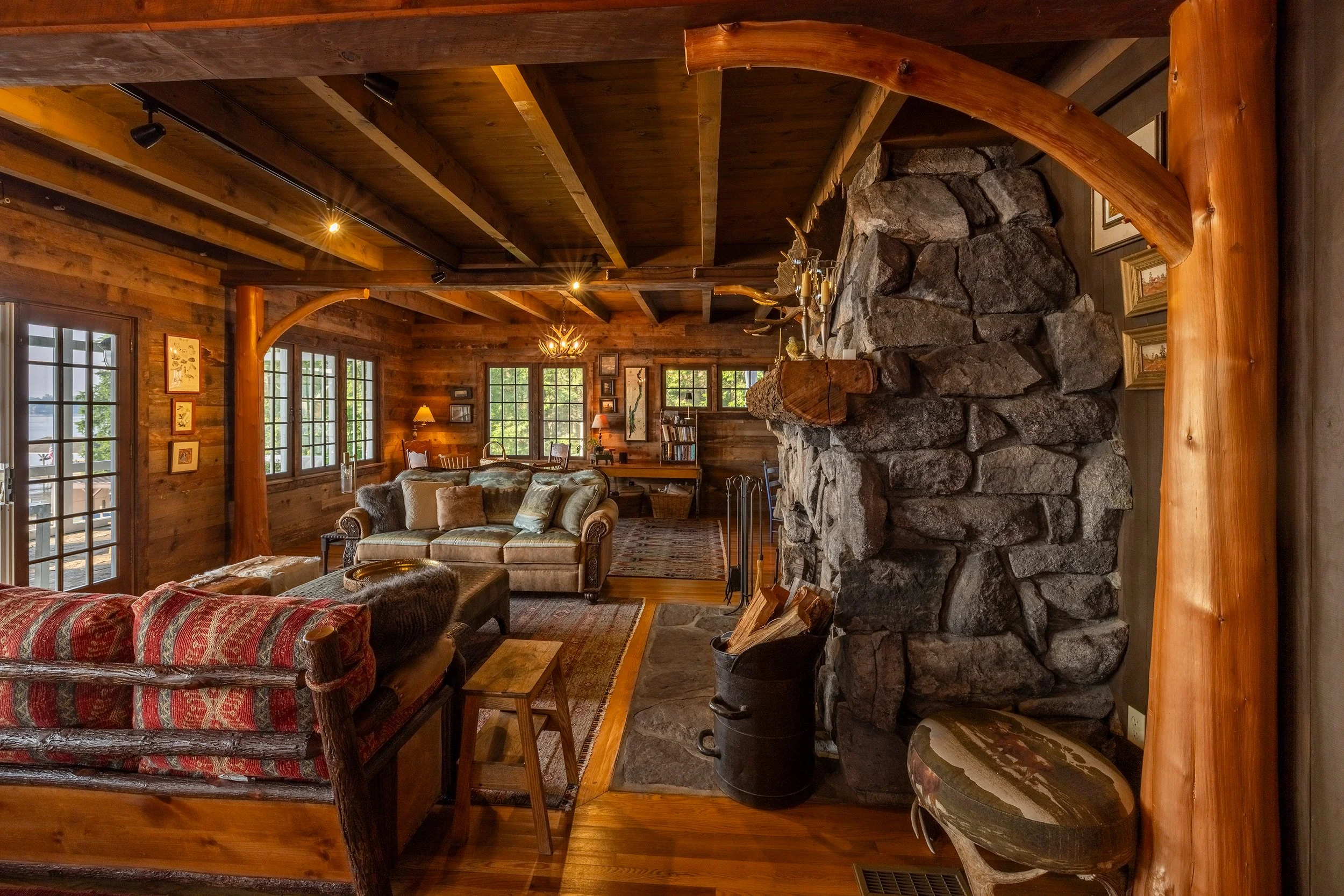 A rustic living room with wood-paneled walls and ceiling, large windows, a stone fireplace, cozy furniture, and warm lighting.