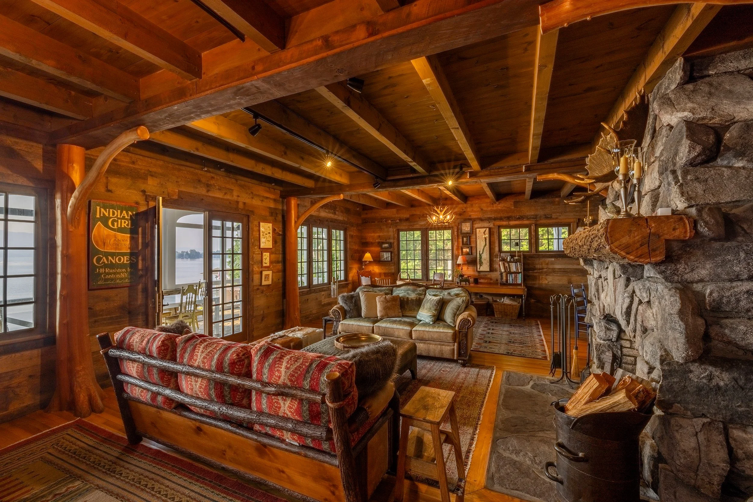 Cozy rustic living room with wood-paneled walls and ceiling, stone fireplace, vintage furniture, and large windows showing a water view.