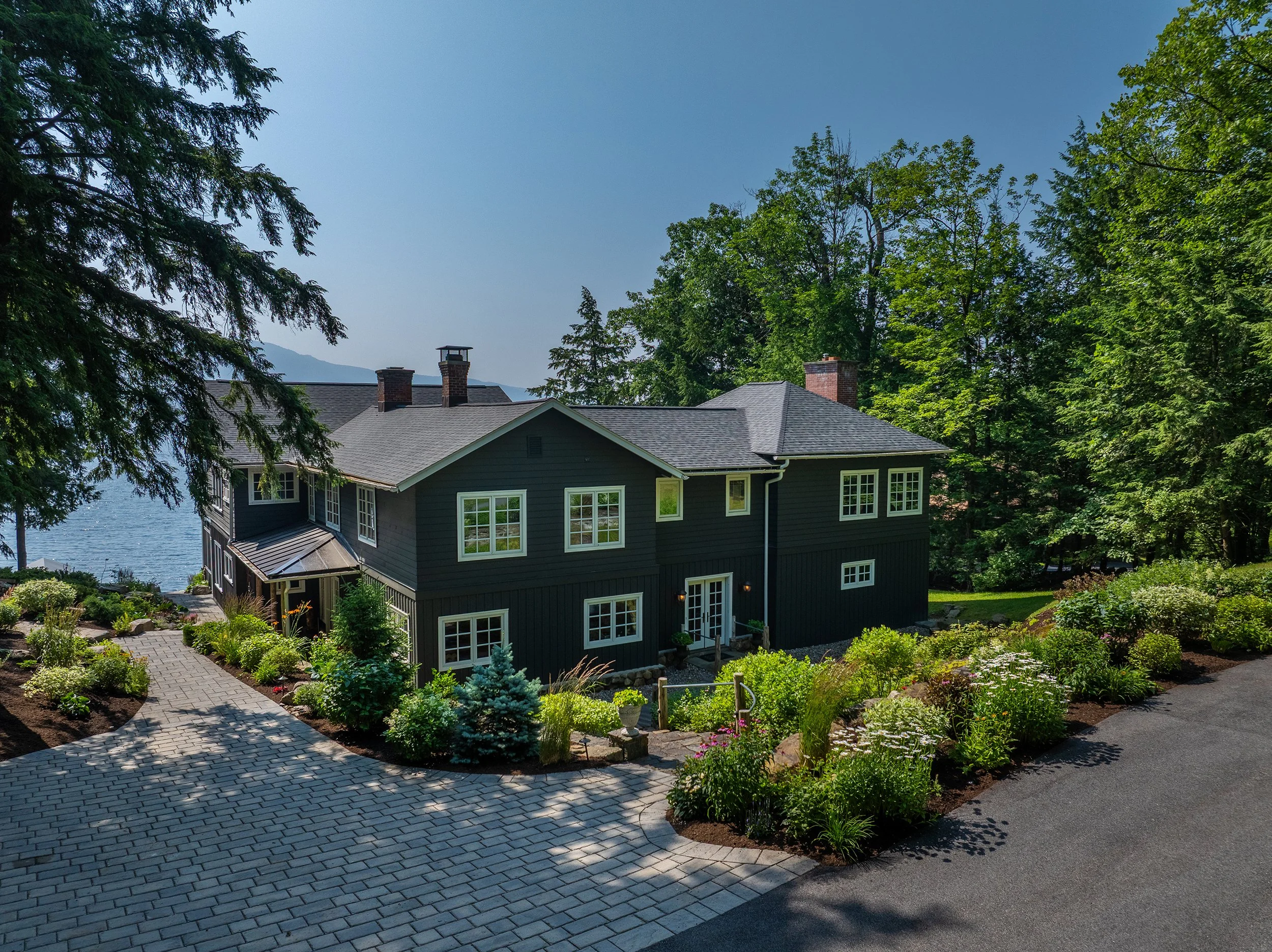 A large dark blue house with white trim, surrounded by lush landscaping and trees, on lakefront with mountains in the background.