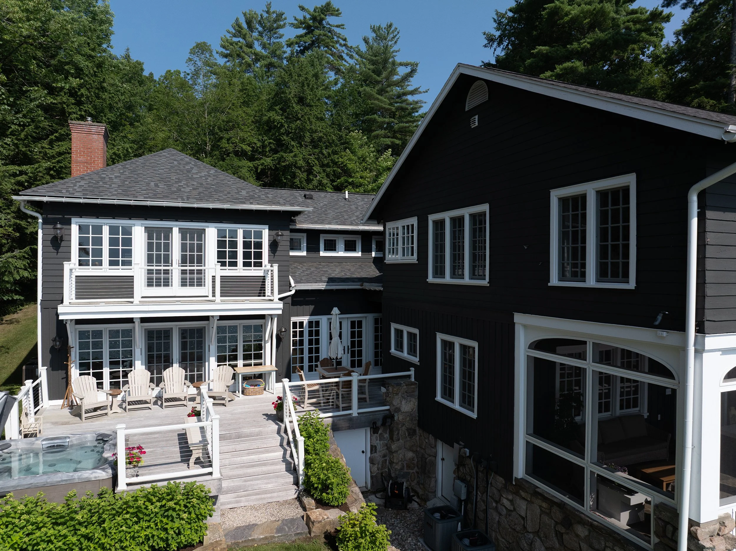View of a dark blue two-story house with multiple white-framed windows, a balcony, a patio with Adirondack chairs, a hot tub, and surrounding greenery and trees.