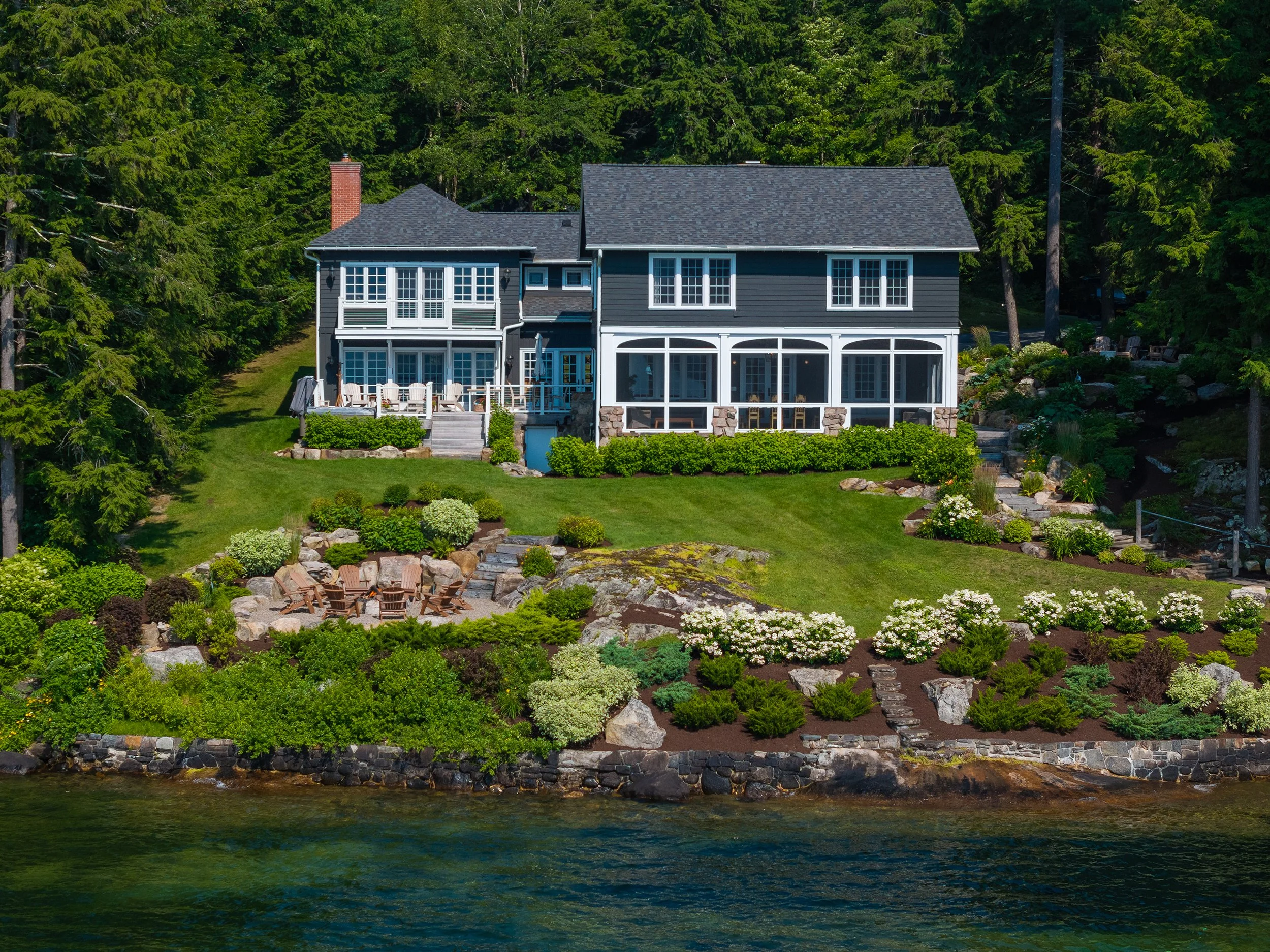A two-story house with dark siding and white trim is situated on a hillside overlooking a body of water. The house features large windows, a screened-in porch, and a deck with outdoor furniture. The landscaped yard has lush green grass, a flower gard