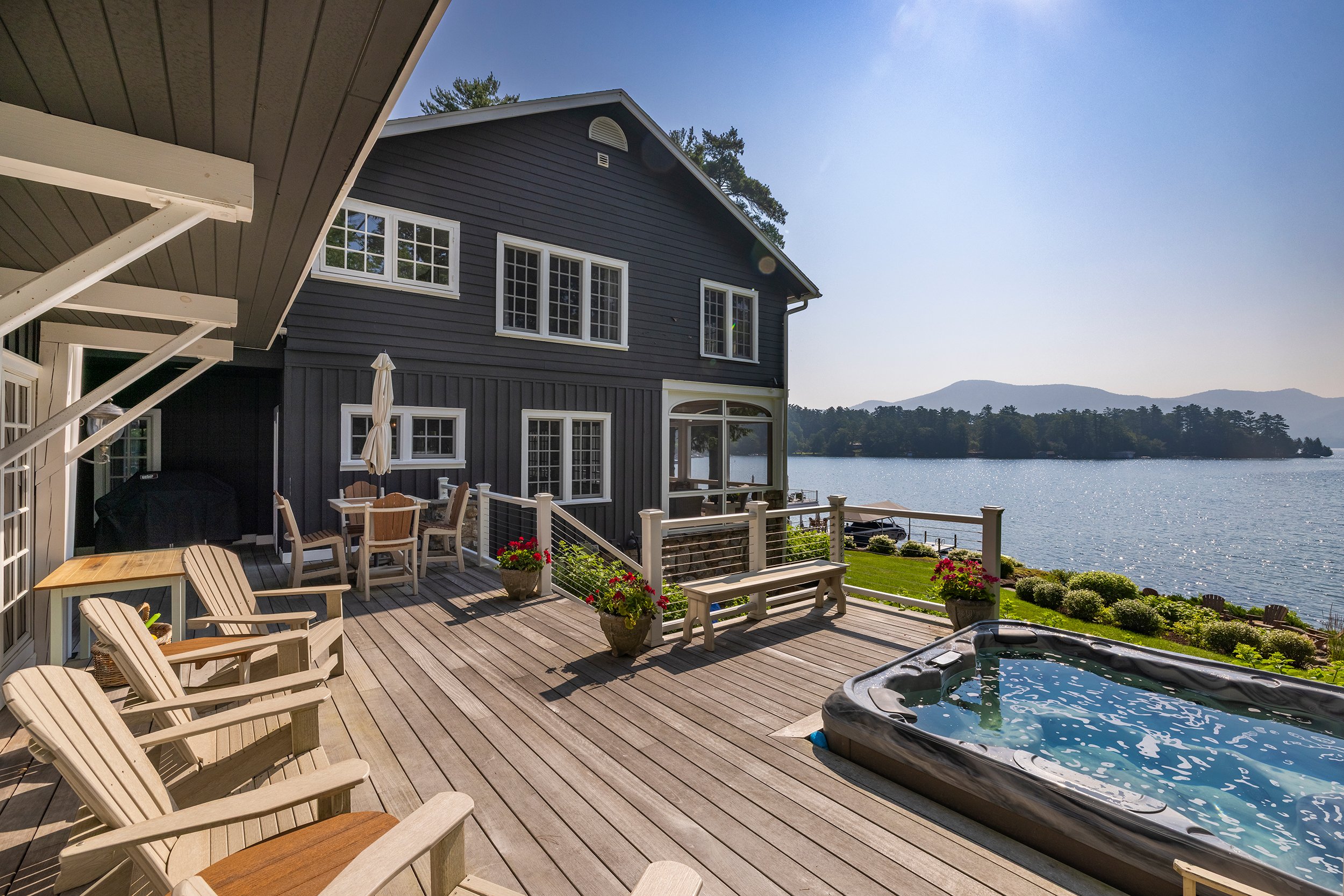A lakeside deck with Adirondack chairs, a hot tub, potted flowers, and outdoor dining furniture overlooking a lake with a house and mountains in the background.