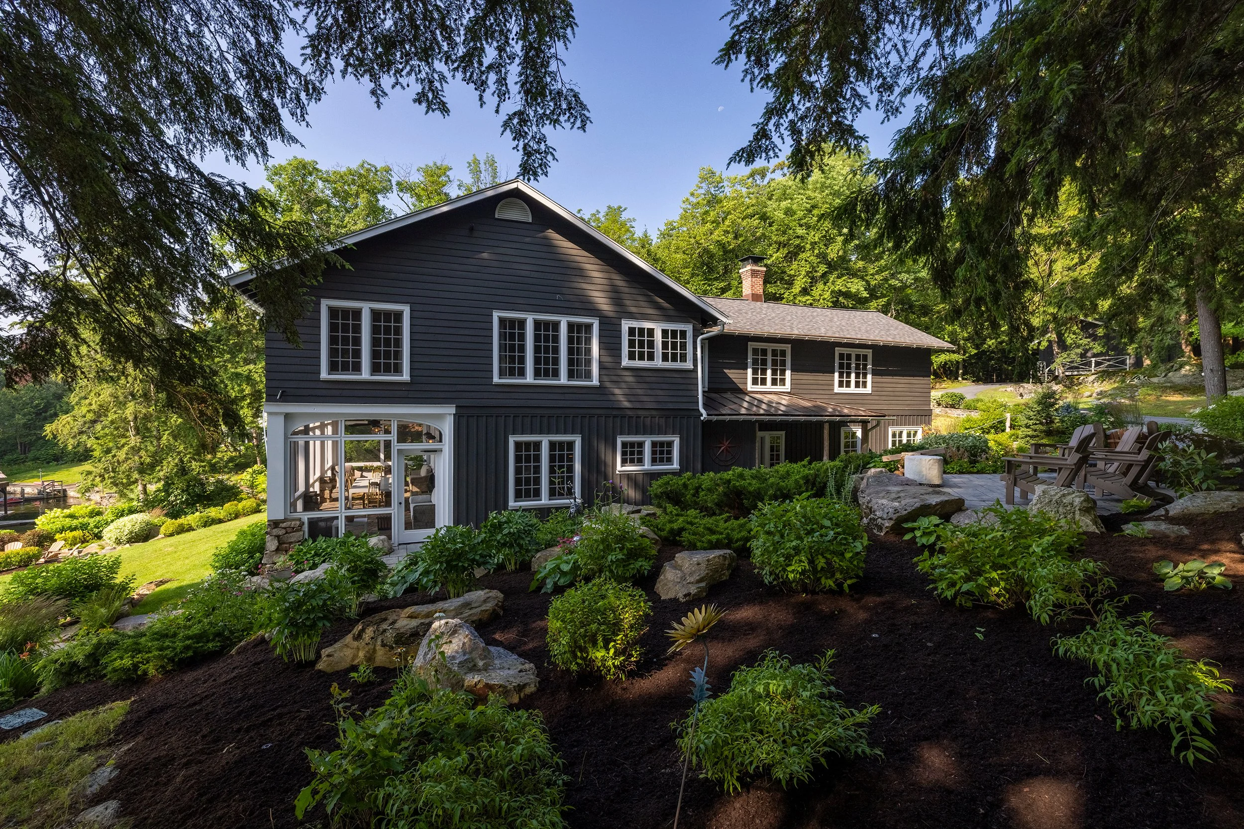 A two-story dark blue house with white window frames surrounded by lush landscaping with rocks and trees, under a clear blue sky.