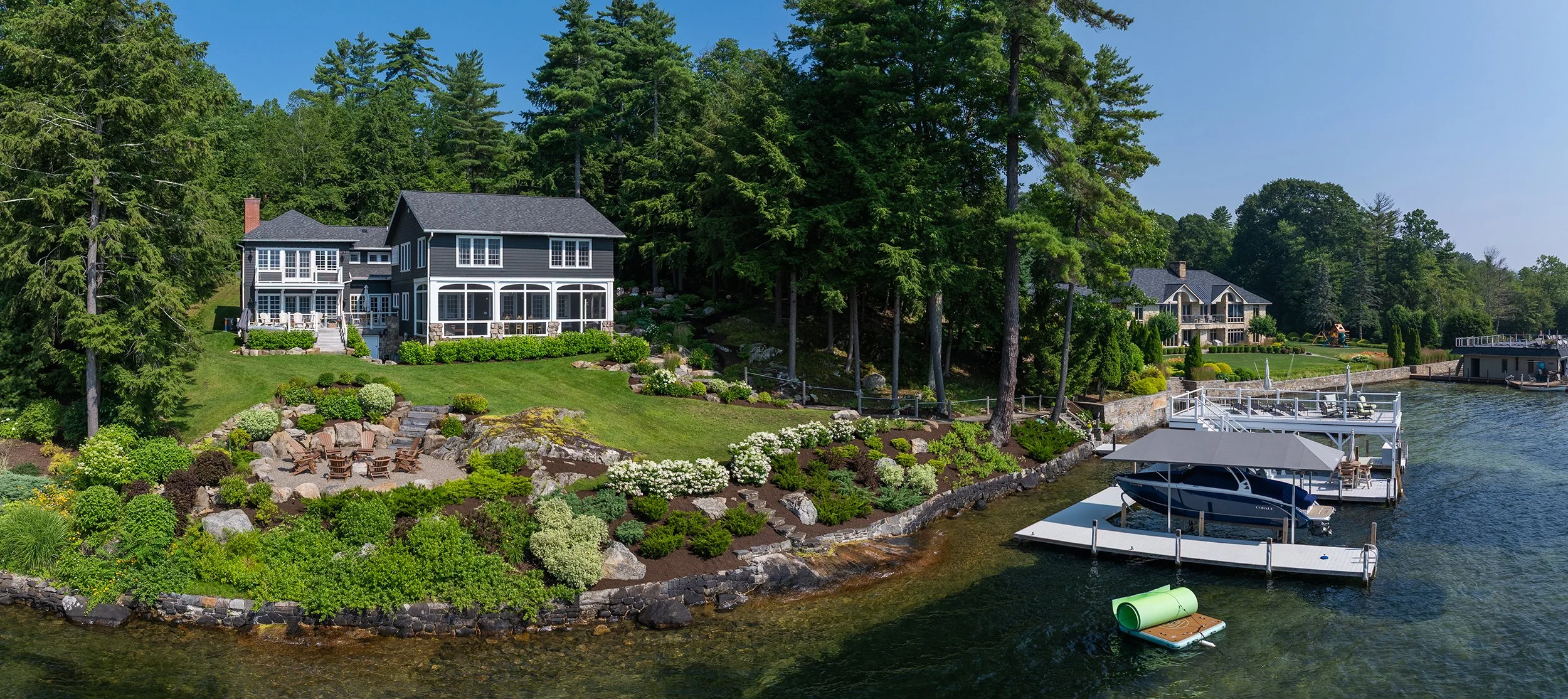Lakeside property with a large, dark blue house, lush landscaping, and a dock with boats and a swimming platform.