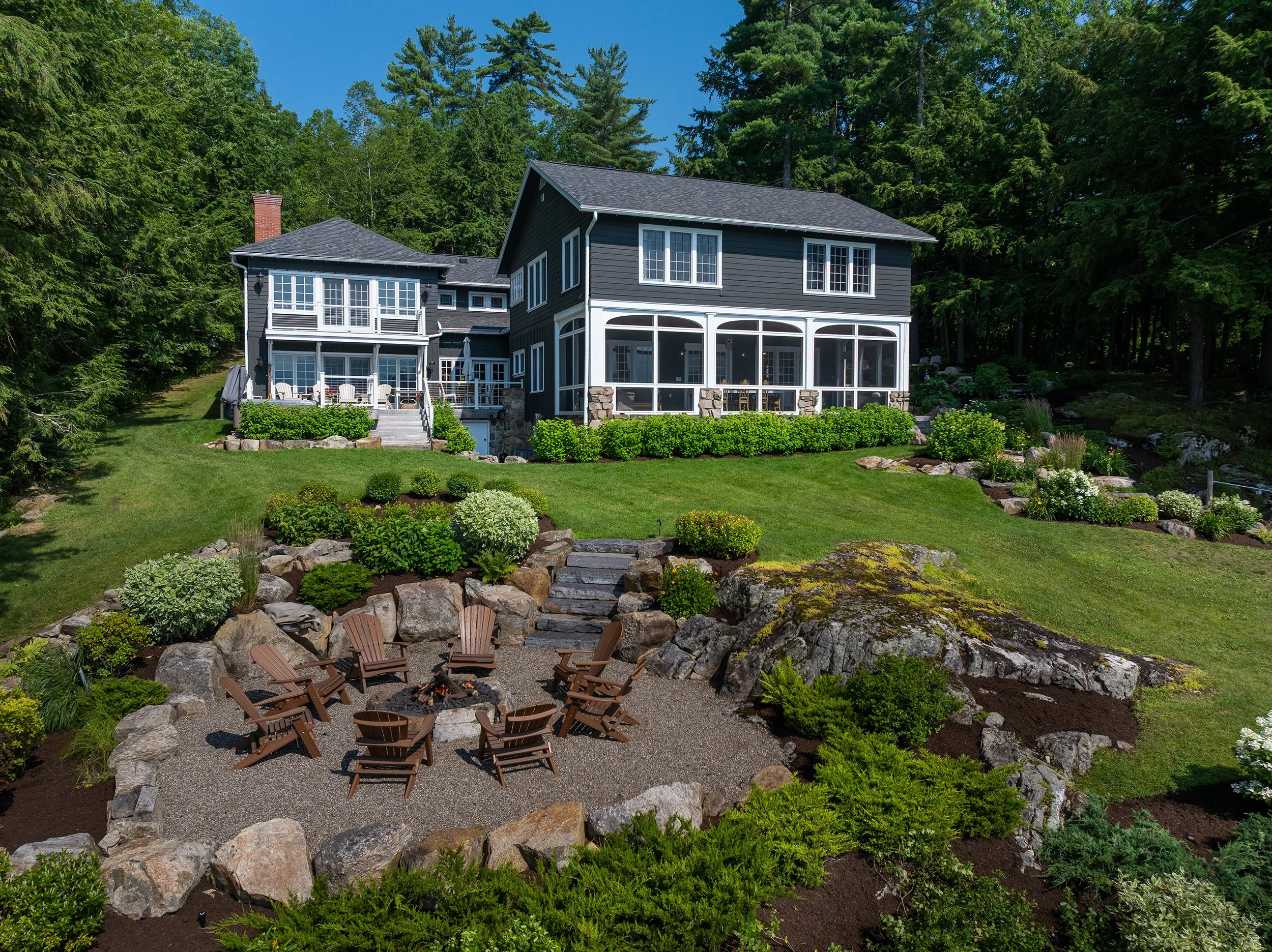 A large, dark blue house with white trim surrounded by lush greenery and landscaping, featuring a backyard with a circular fire pit area with wooden Adirondack chairs and a stone patio with outdoor furniture, set in a wooded area on a sunny day.