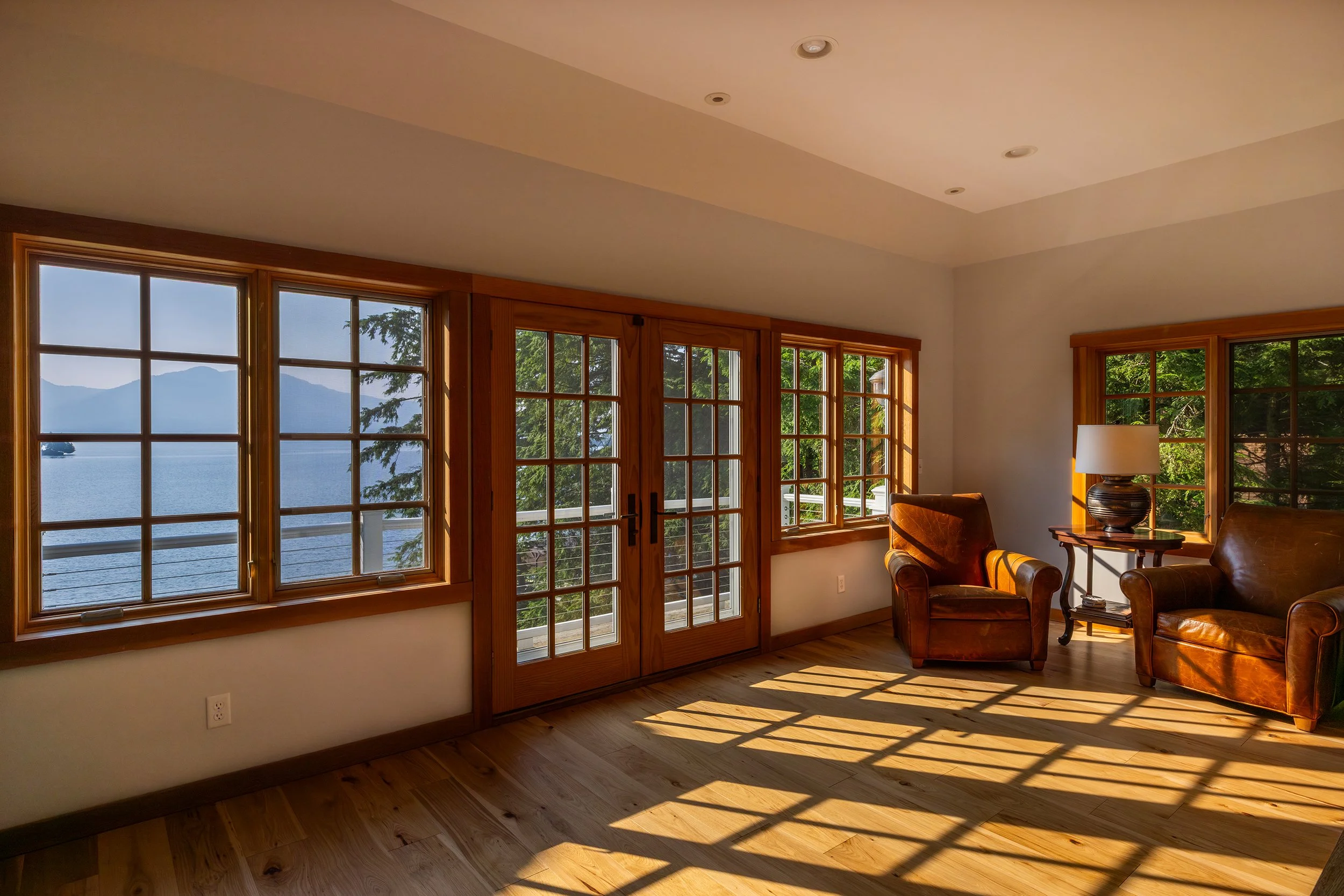 A cozy sitting room with large windows and a glass door, revealing a view of a lake and mountains outside. The room features two leather armchairs, a side table with a lamp, and sunlight casting shadows on a hardwood floor.