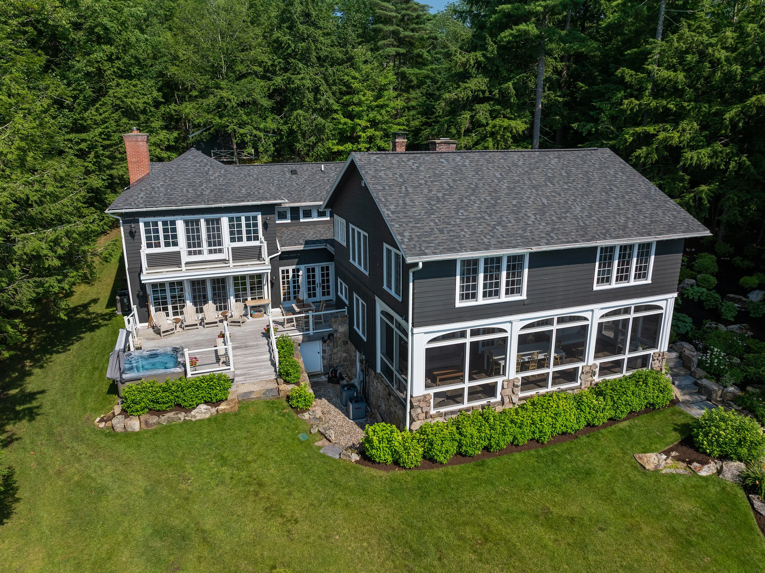 A two-story house with dark blue siding, white trim, and a multi-pitched roof surrounded by a green lawn and trees, featuring a screened porch and an outdoor deck with furniture and a hot tub.