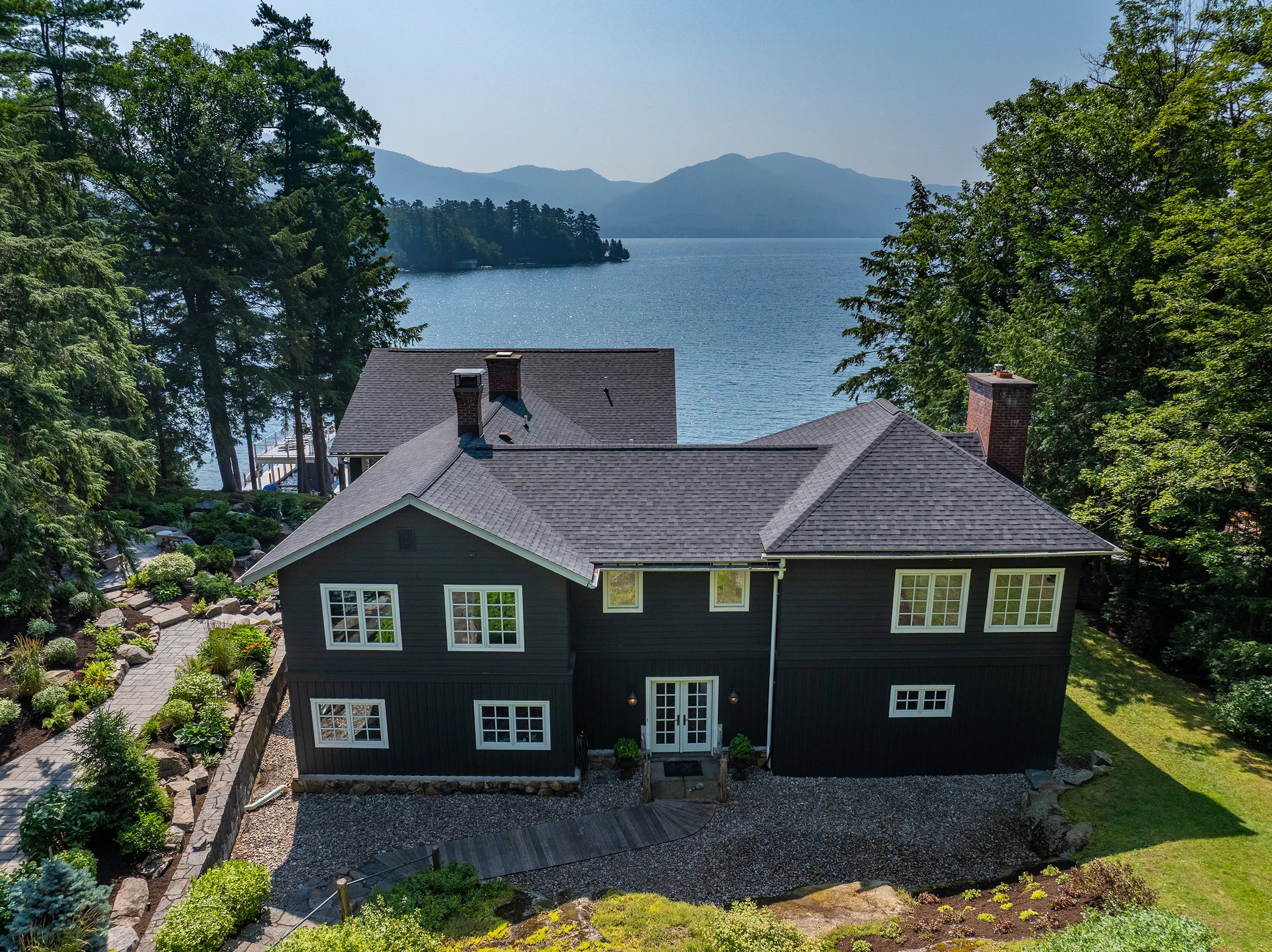 A two-story dark blue house with a grey shingle roof, surrounded by trees and a well-maintained garden, overlooking a lake with mountains in the background.