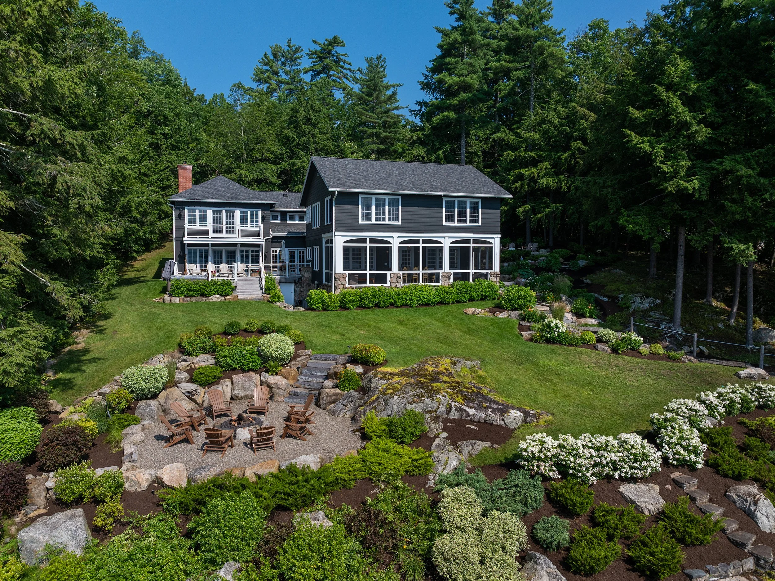 A large, dark blue house situated on a lush green, elevated lawn with landscaped garden beds, patio, and a fire pit area with Adirondack chairs, surrounded by tall trees under a clear blue sky.
