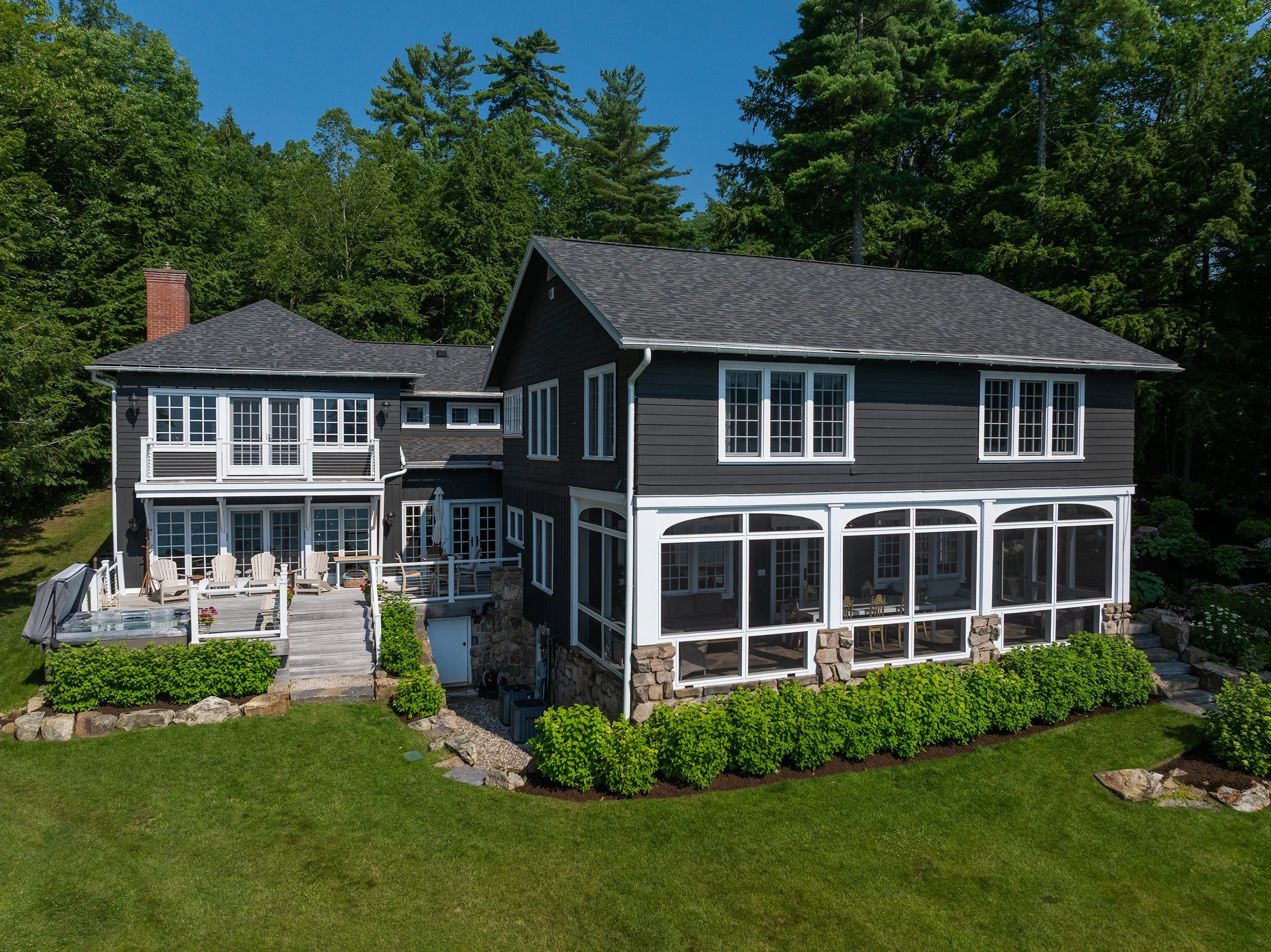A large dark blue house with multiple windows and a screened porch, surrounded by green grass, shrubs, and trees under a clear blue sky.