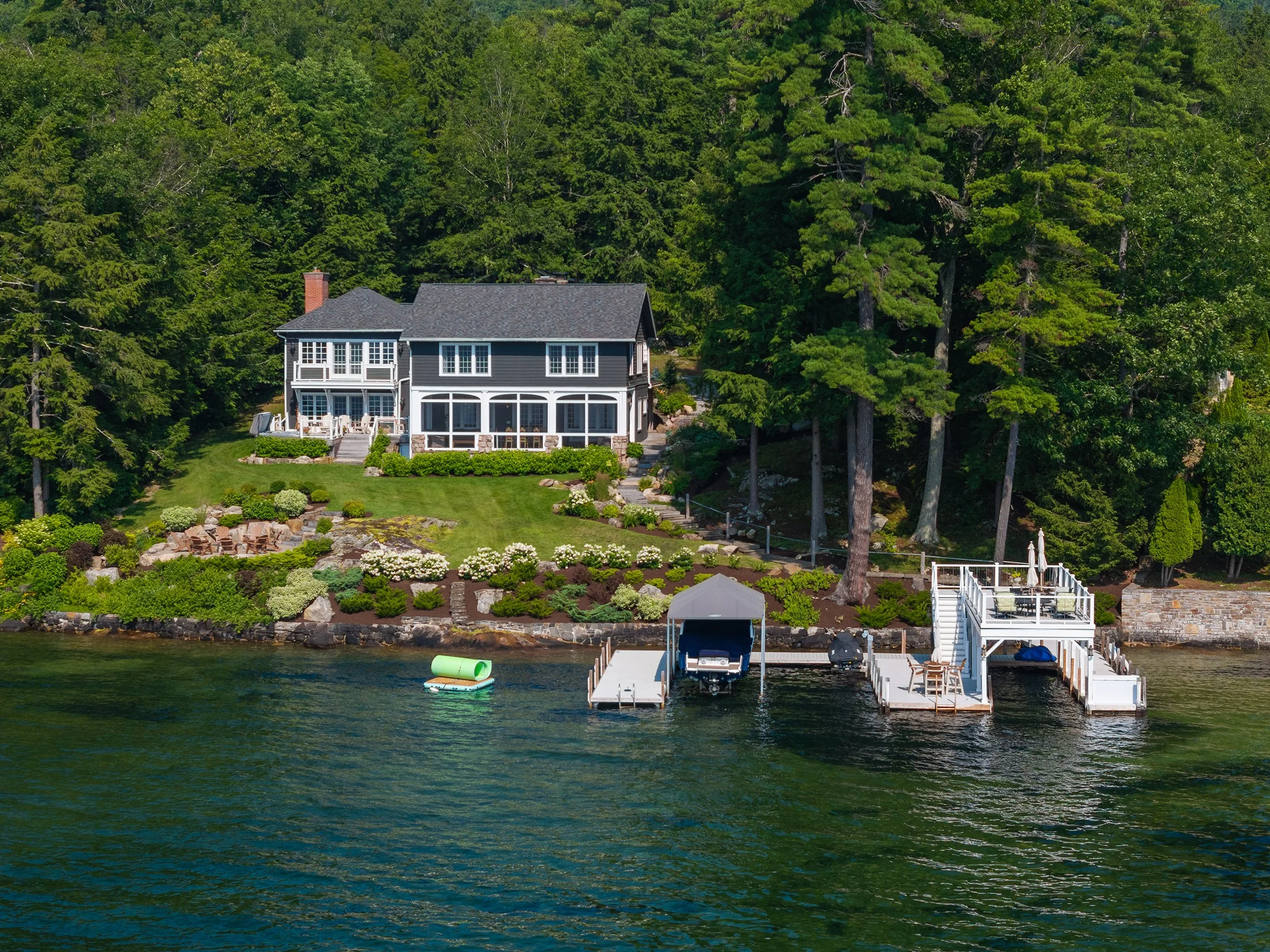 A large waterfront house with a lush garden, trees, and a boat dock on a lake.