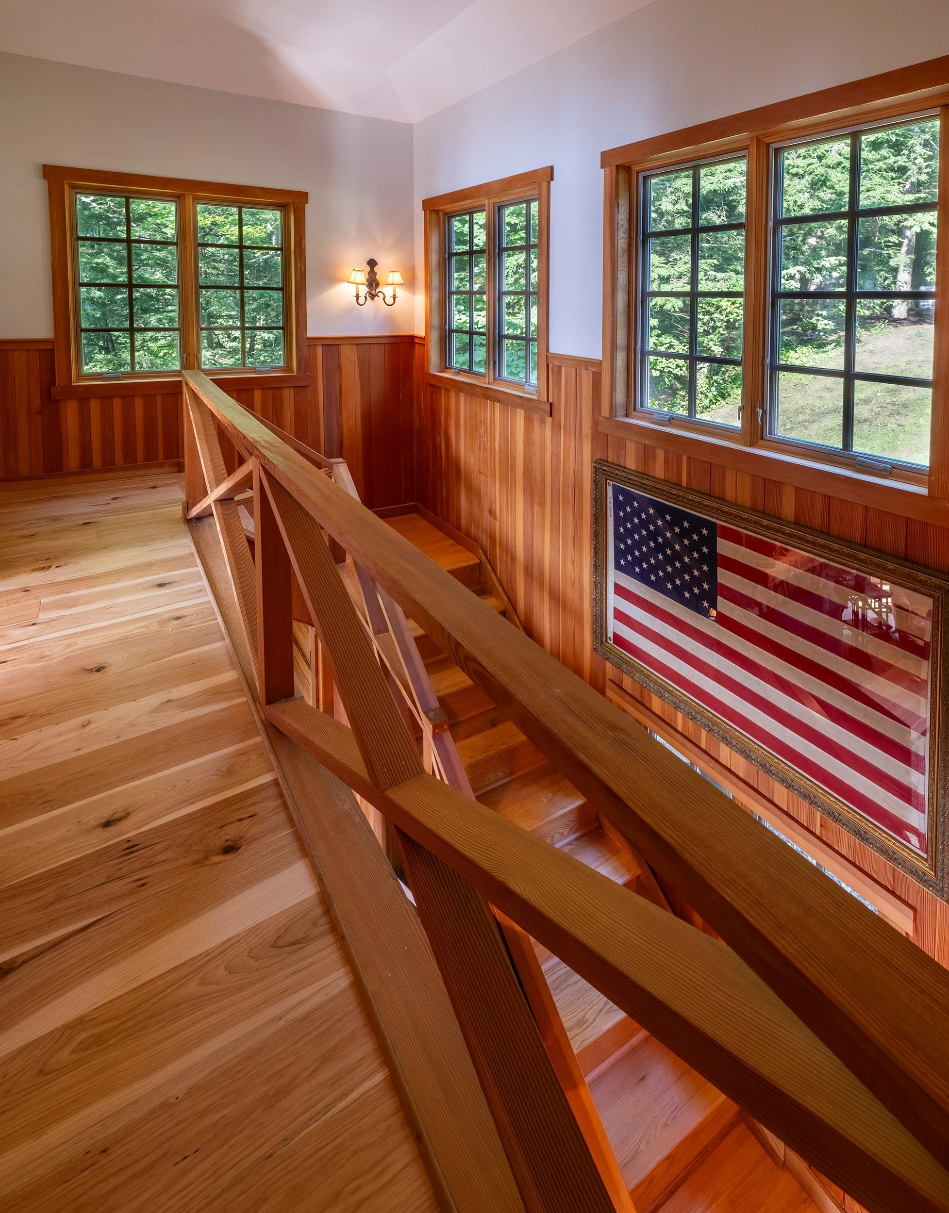 Wooden staircase landing with windows overlooking greenery, American flag in frame, warm wall-mounted light fixture.