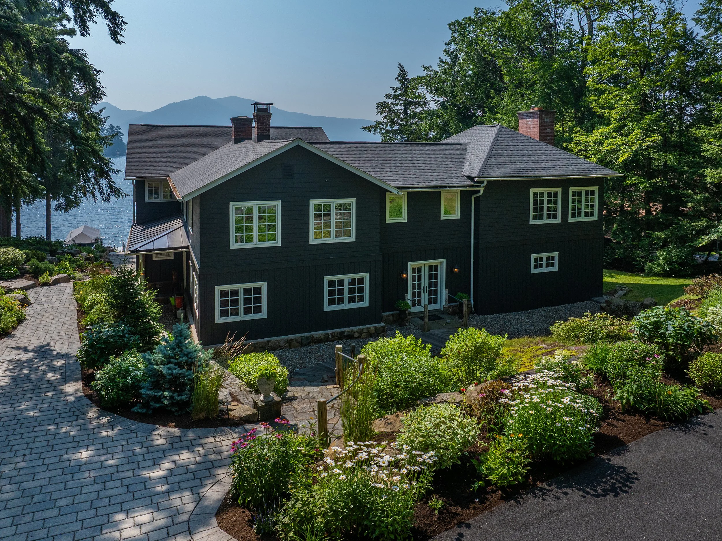 A dark blue two-story house with white window frames, surrounded by a lush green garden, with a lake and mountains in the background.