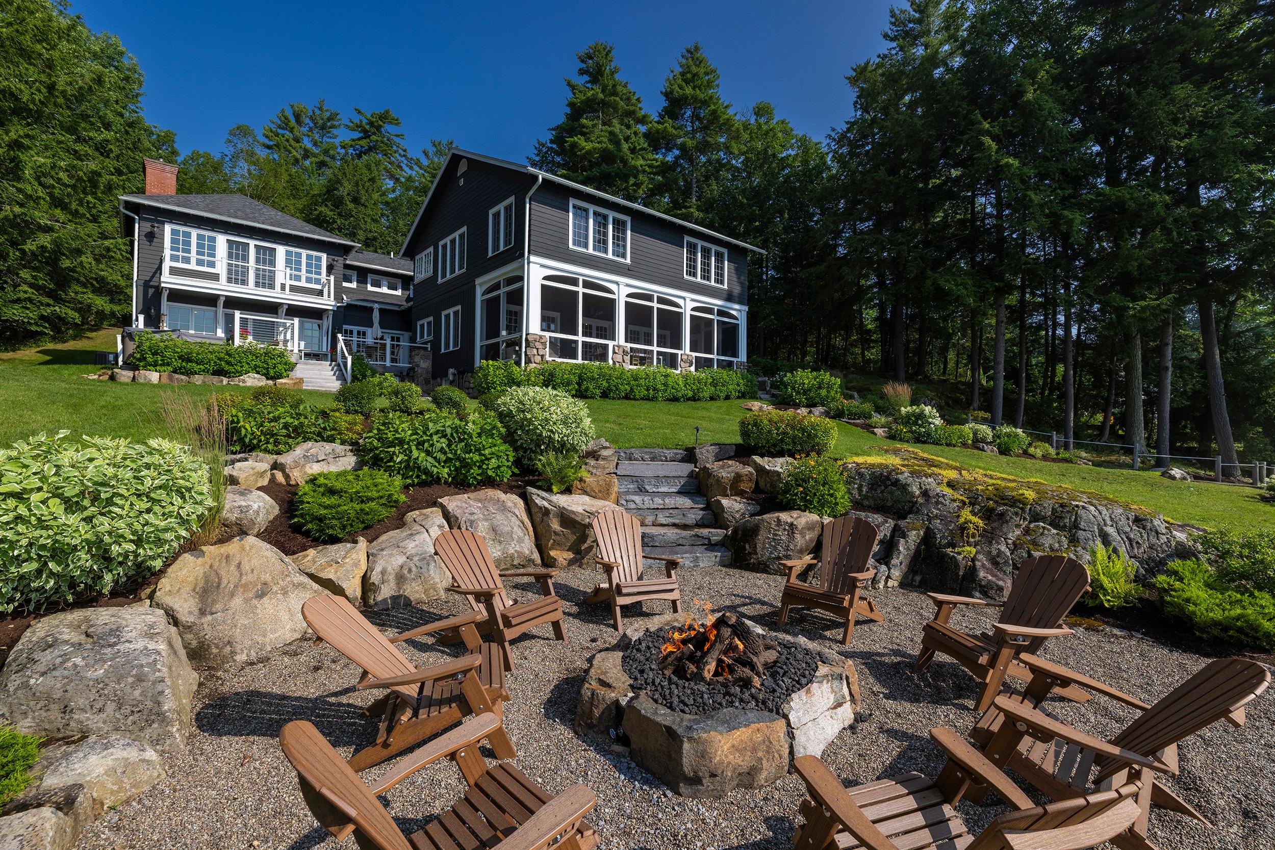 A backyard with a fire pit surrounded by wooden Adirondack chairs, stone steps, landscaped bushes, and a large dark blue house with multiple levels and a screened porch, set against a backdrop of dense trees under a clear blue sky.