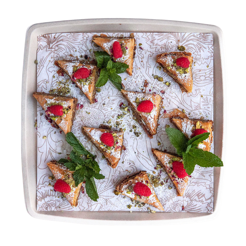 A white tray with triangular slices of dessert topped with raspberries, mint leaves, powdered sugar, and chopped pistachios.